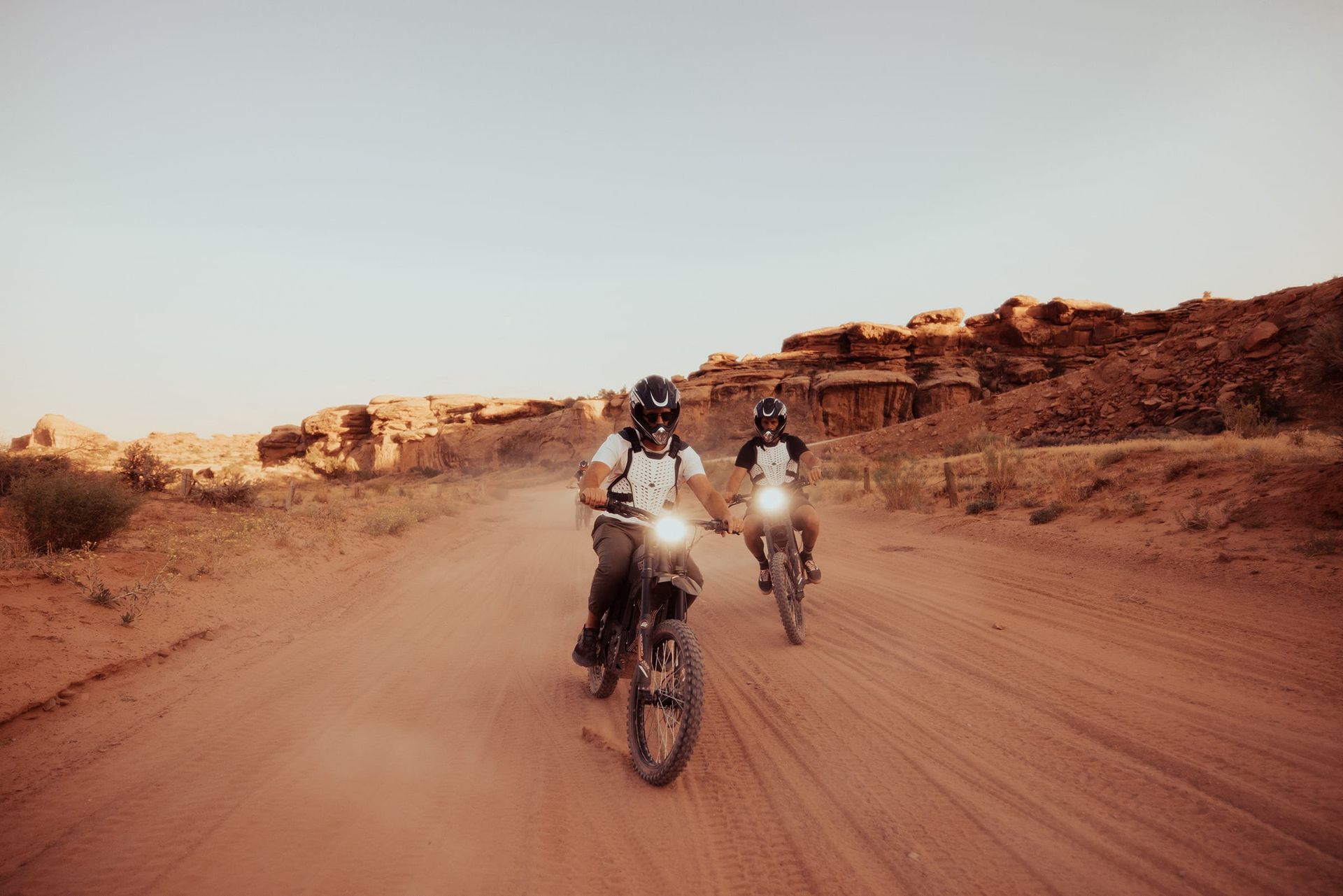 Two people ride motorcycles on a dusty road in a desert landscape.