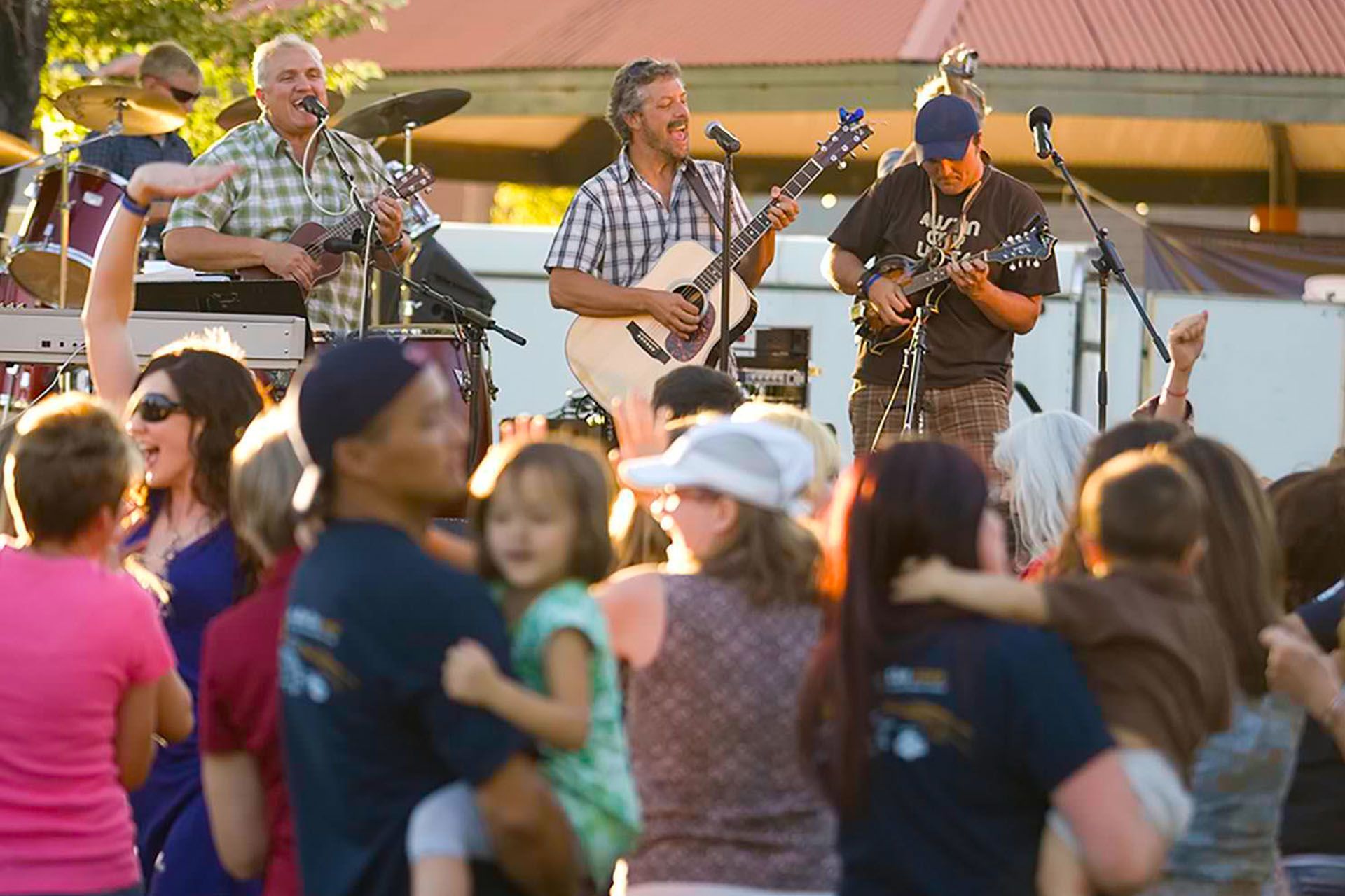 Band performing on outdoor stage; crowd of people dancing. Golden hour sunlight.