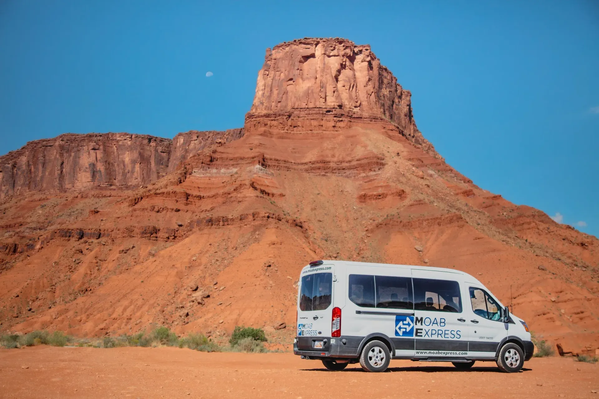 White shuttle van parked in front of a large red rock formation under a blue sky.