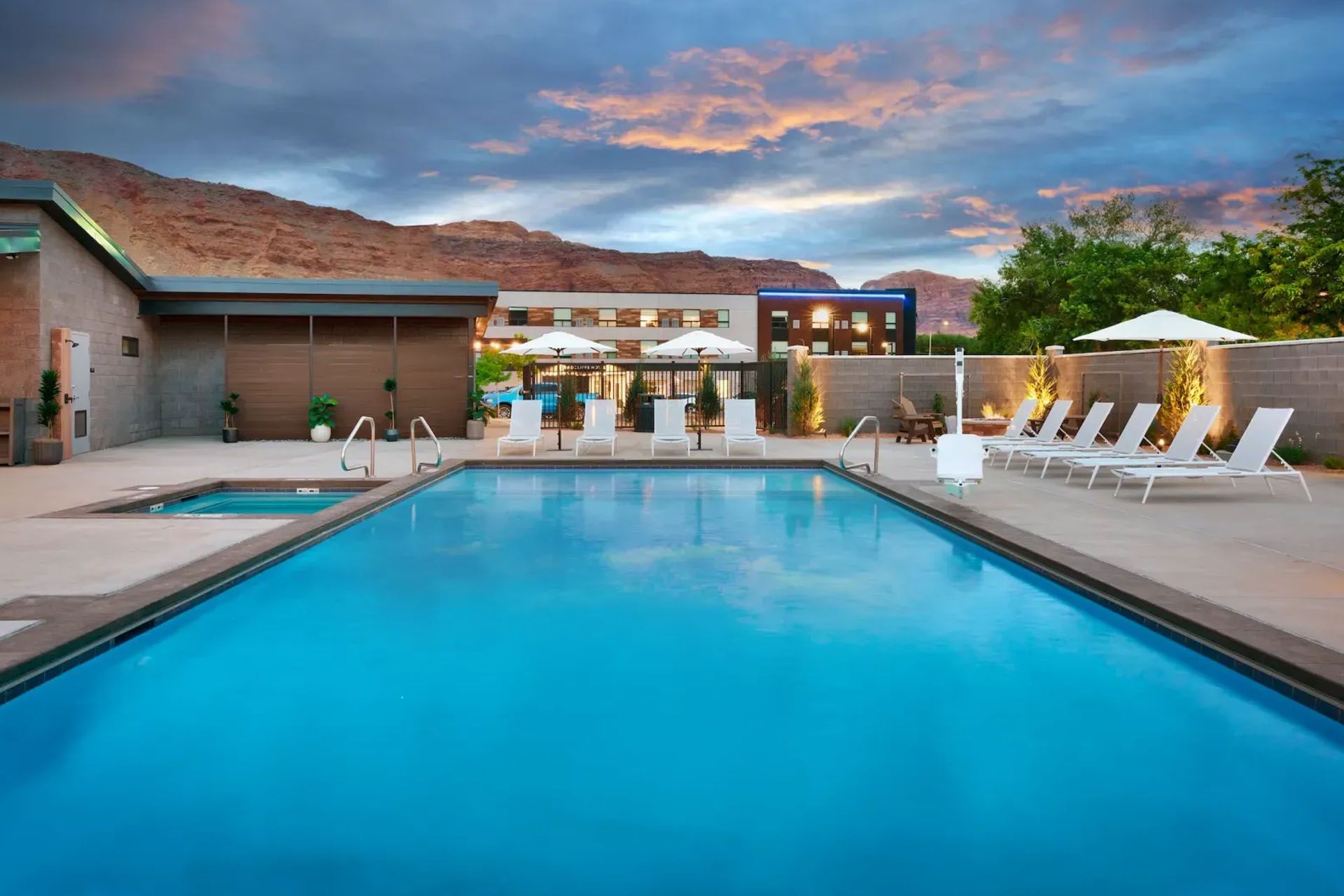 Pool at dusk with mountains in the background, lounge chairs, umbrellas, and a modern building.