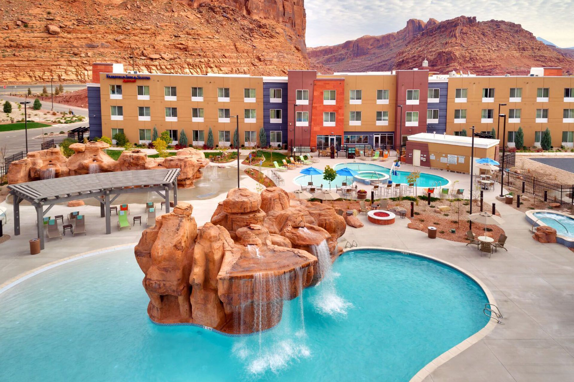 Hotel with colorful exterior, multiple pools, water features, and red rock backdrop.