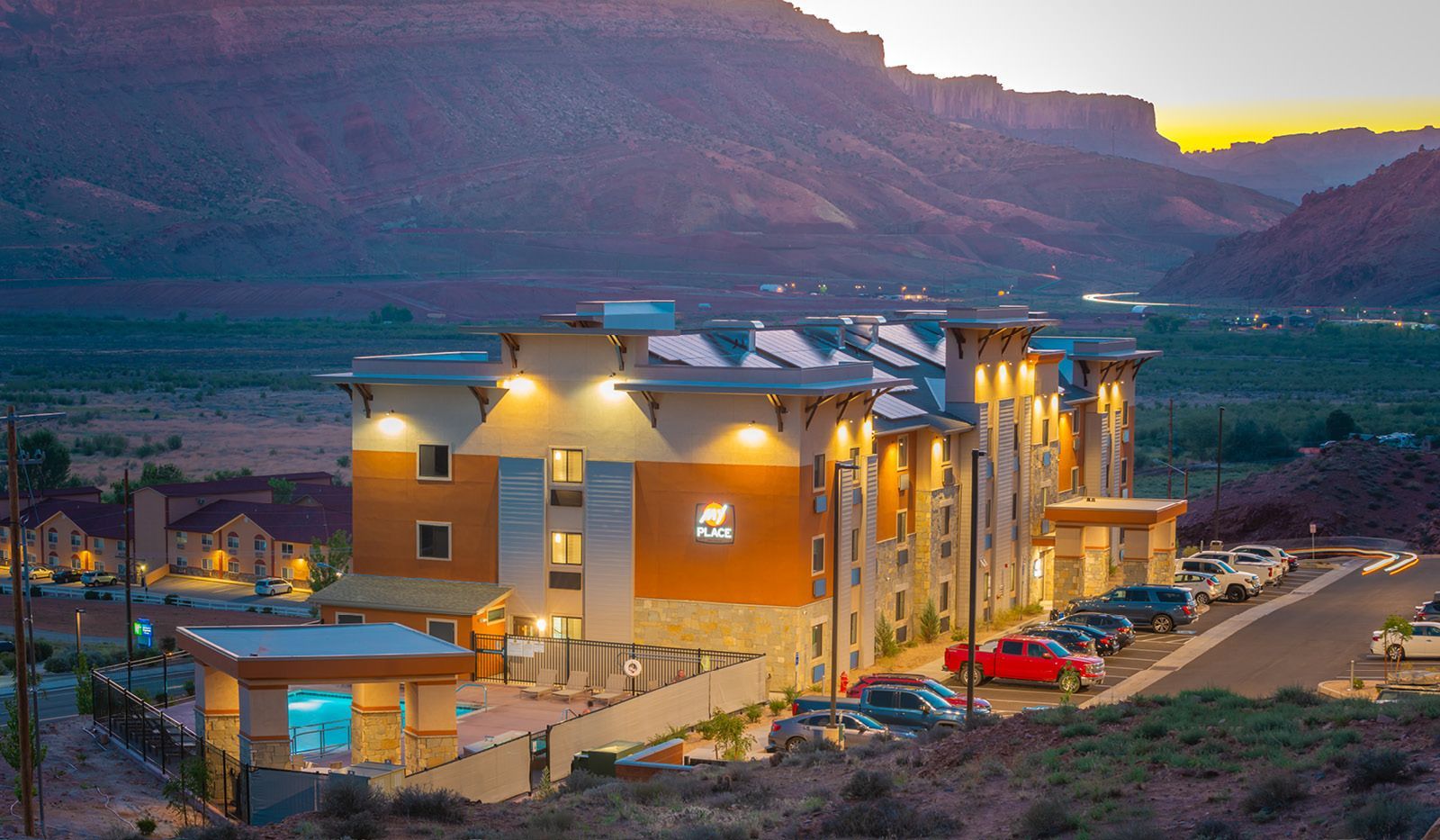 Hotel building in desert landscape at dusk, with lit windows, pool, and parked cars.