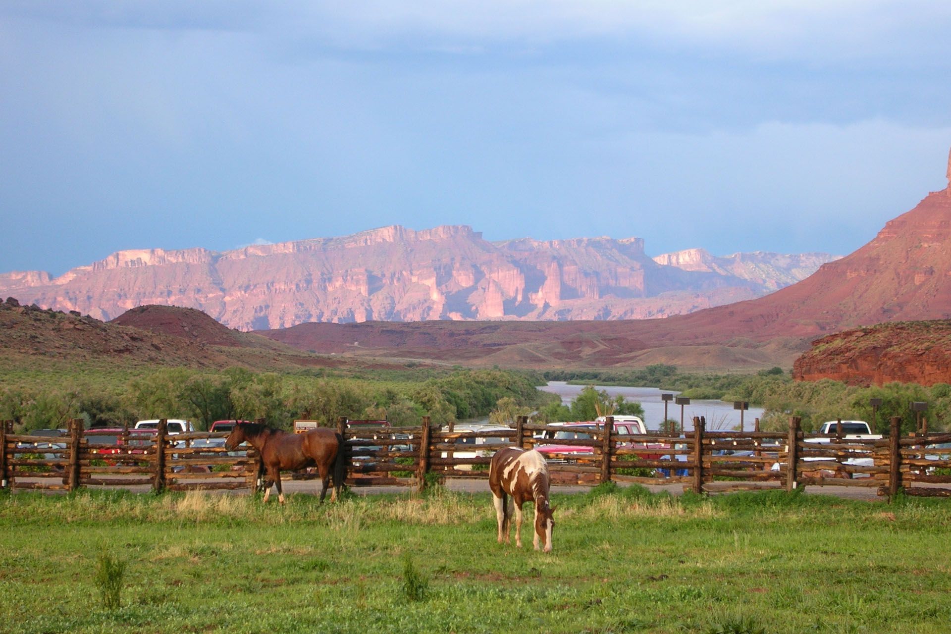Horses in a green field near a wooden fence, with red rock mountains and a river in the background.