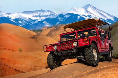 A Hummer vehicle drives across a sandstone fin with the La Sal Mountains in the background.