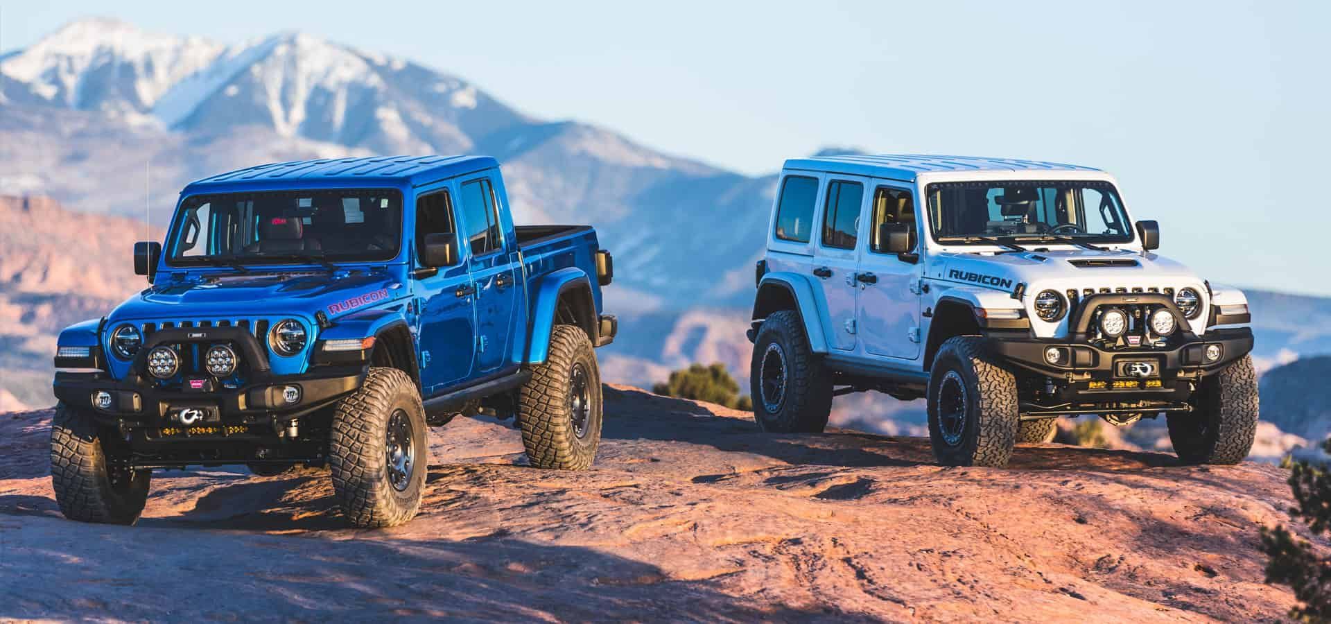 Two Jeeps on a rocky trail, one blue pickup and one white SUV, against a mountain backdrop.