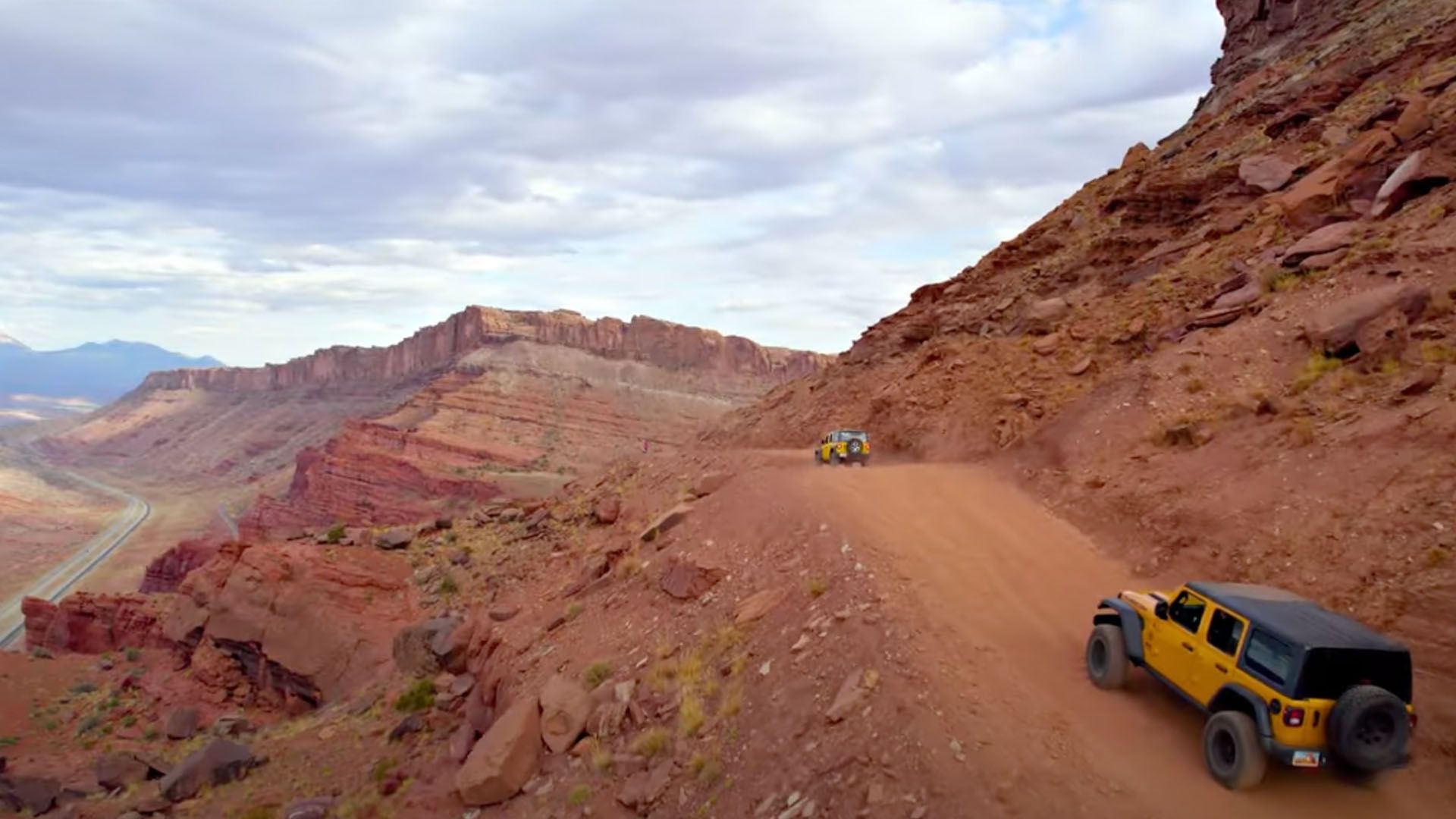 Yellow Jeeps driving along a red dirt road on a mountain side, with red rock formations in background.