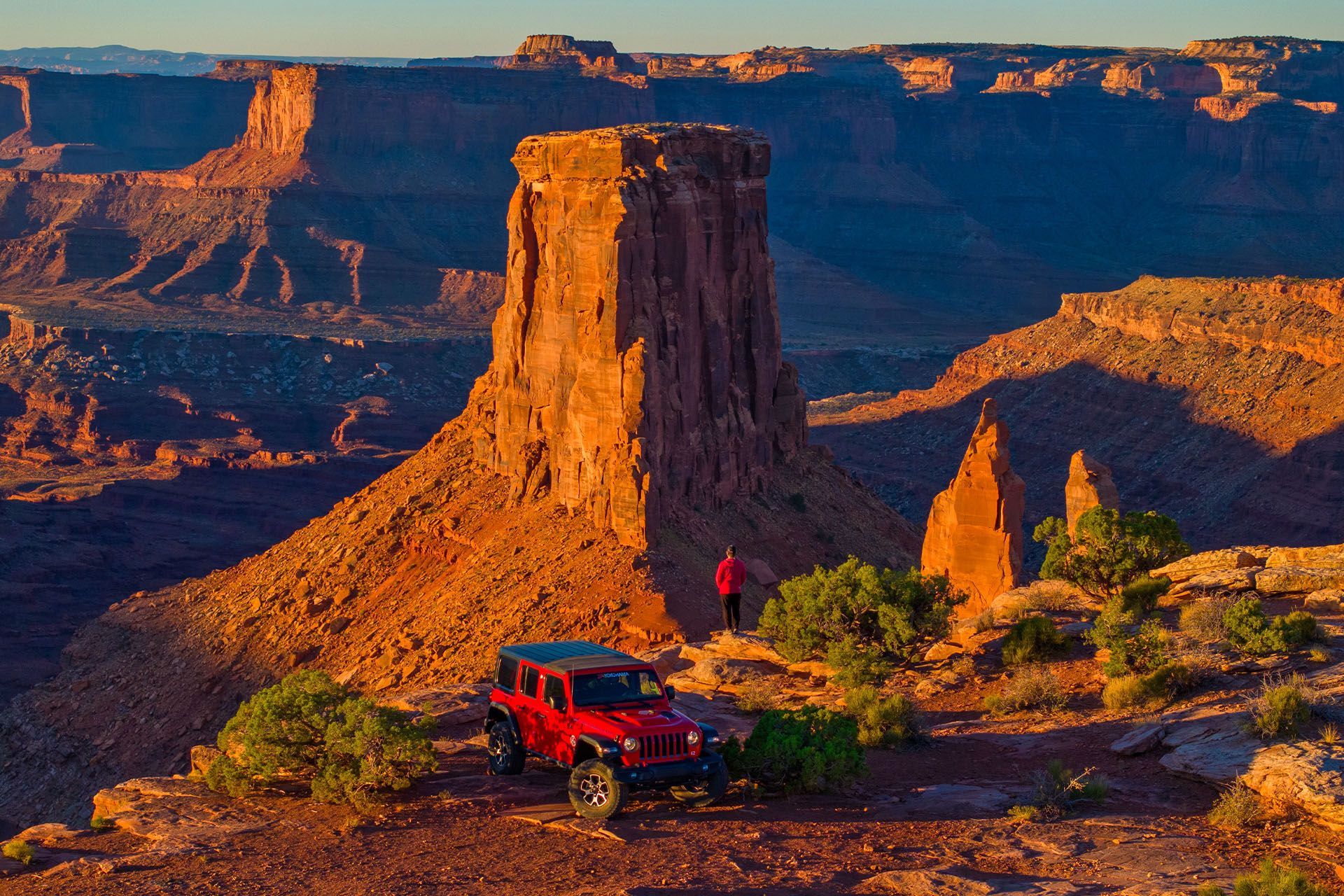 Red Jeep at the edge of a dramatic canyon scene with towering sandstone buttes.