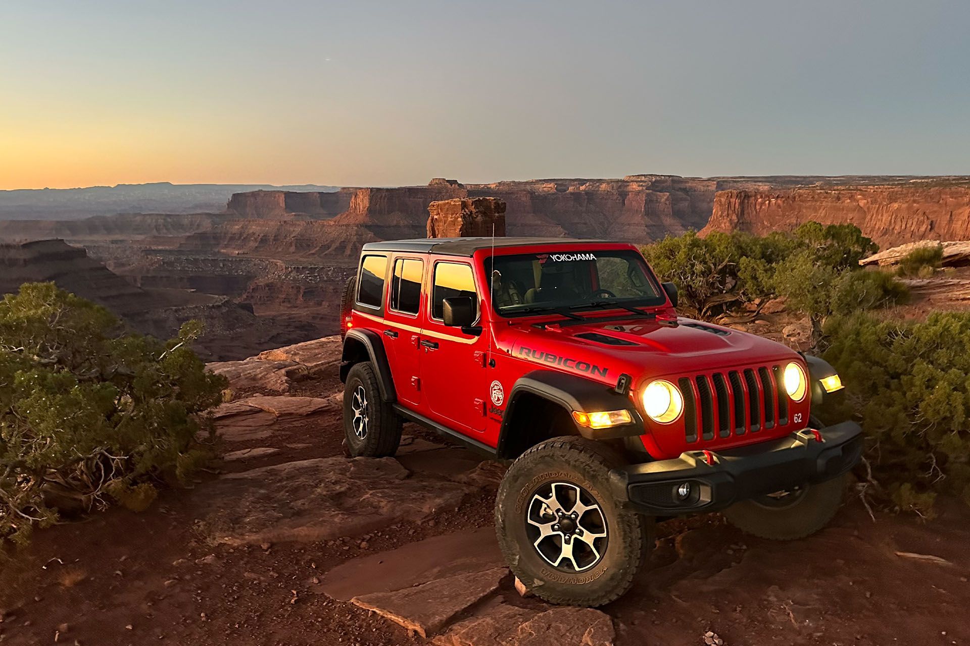 A red jeep rental is parked atop a cliff with a stunning vista behind it.