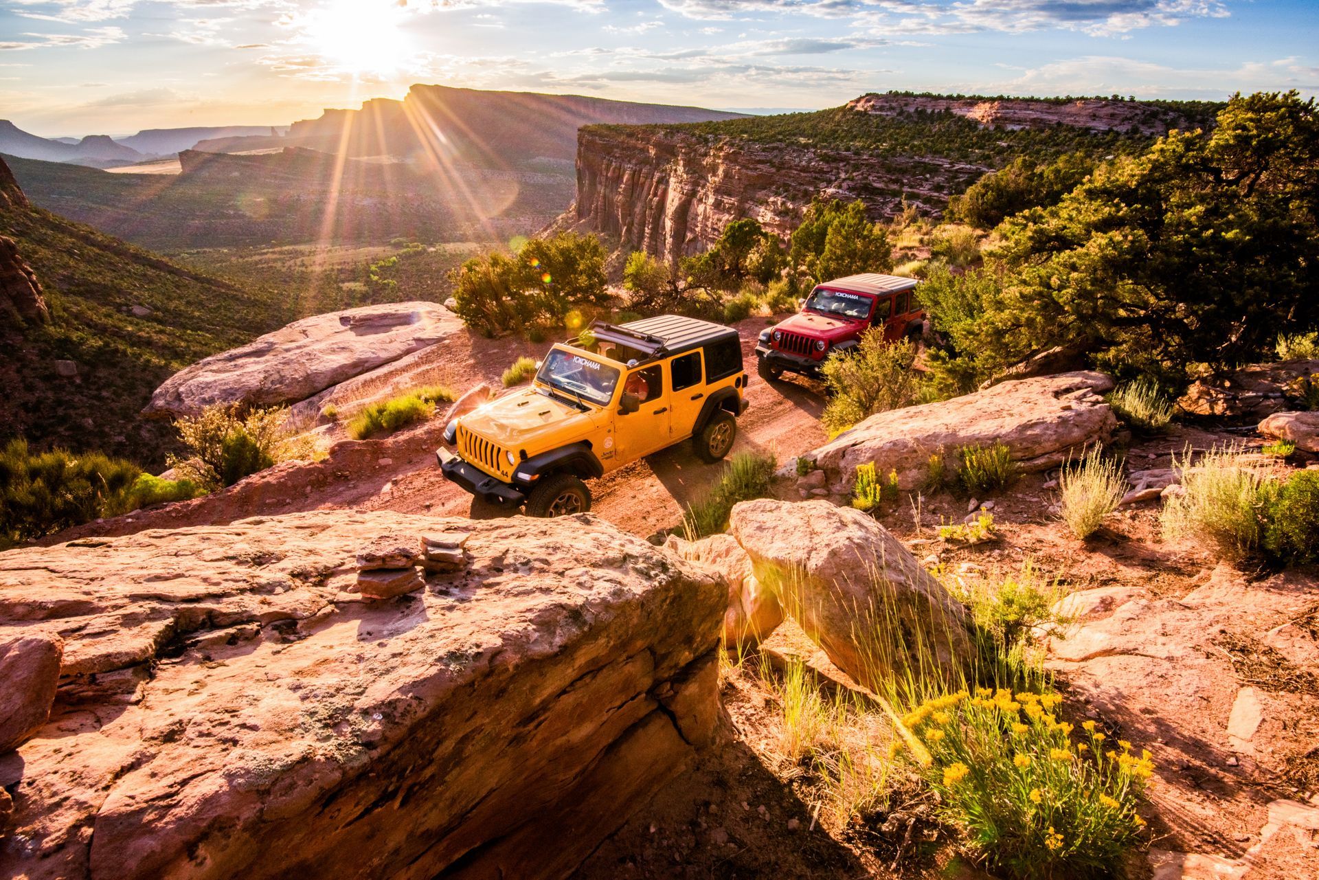 Yellow and red Jeeps driving off-road on a rocky trail with a canyon view at sunset.