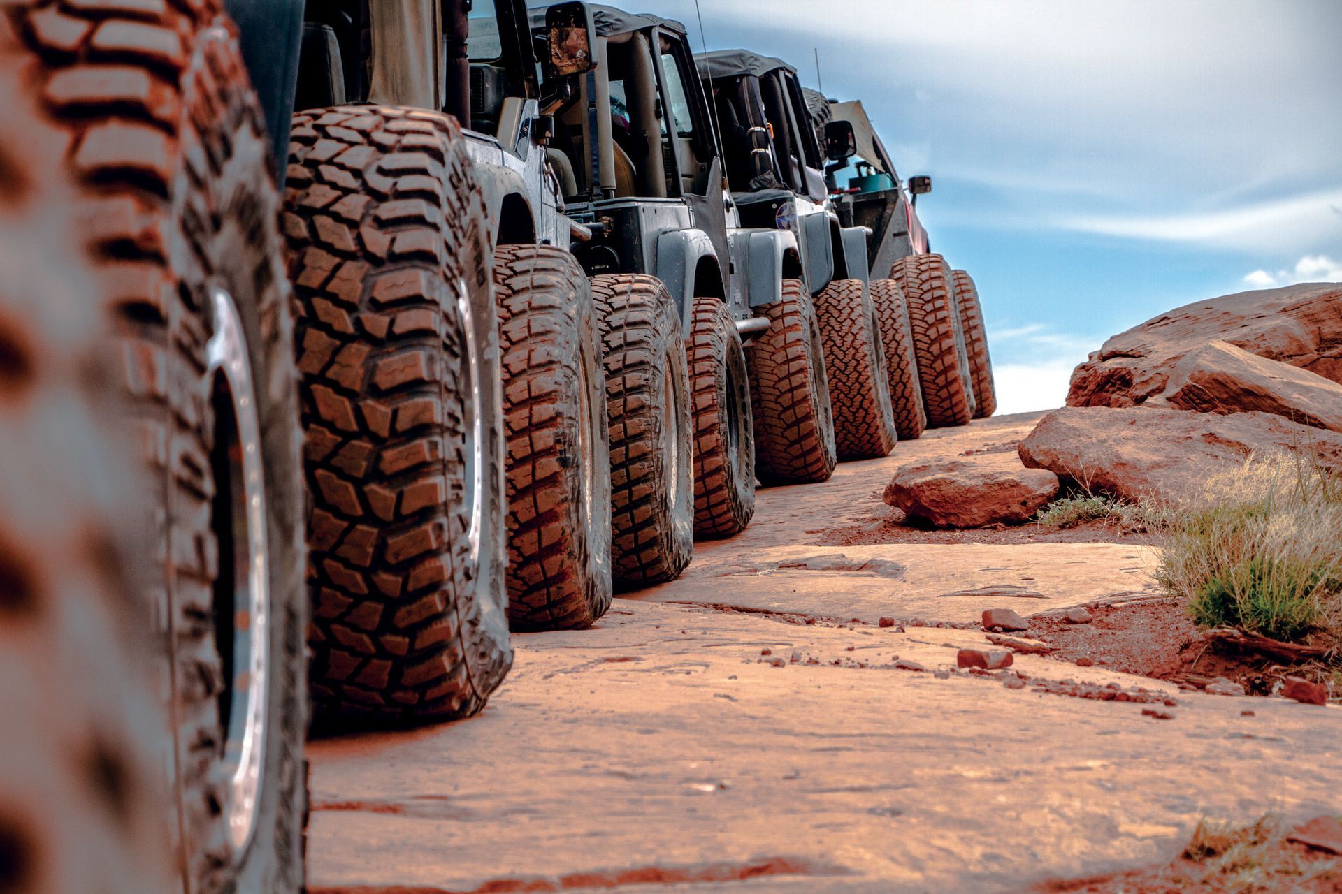 Line of Jeeps with large tires on a red dirt road. Blue sky and rocks in the background.