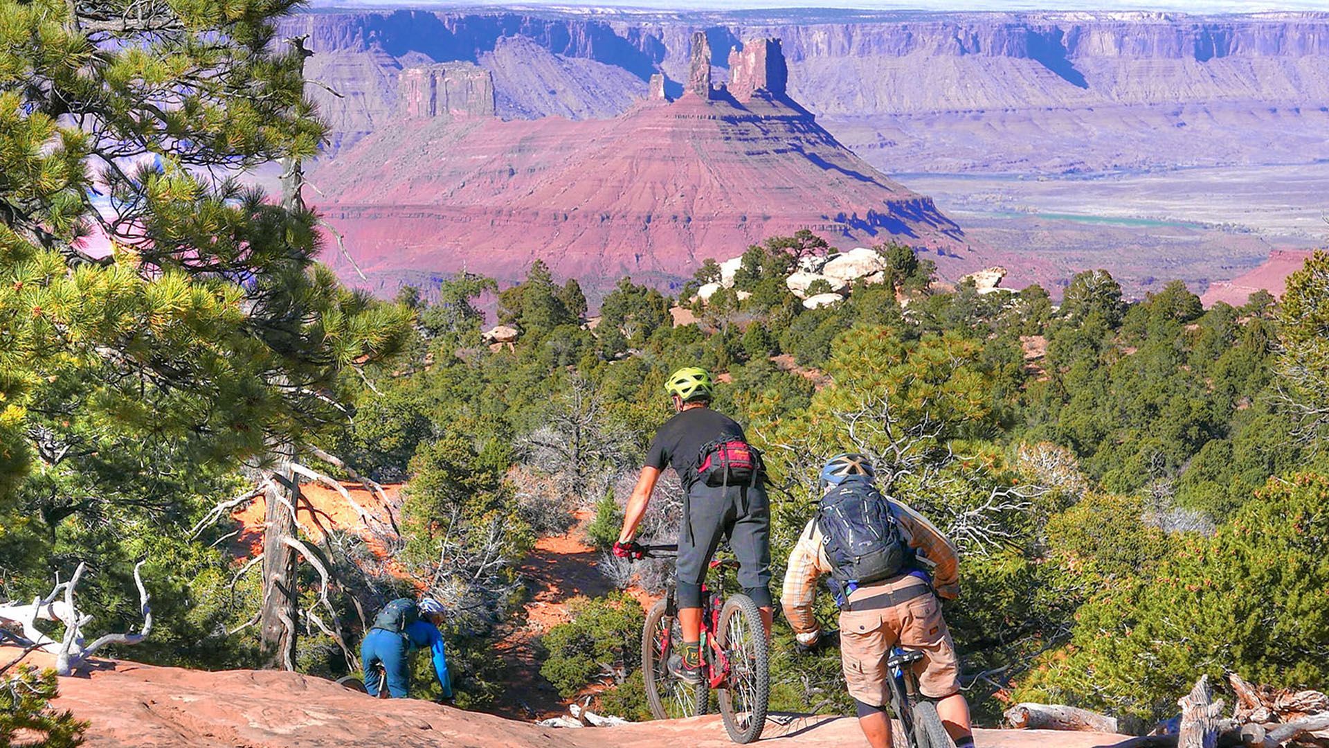 Mountain bikers on a trail overlooking a red rock desert landscape, with blue sky.
