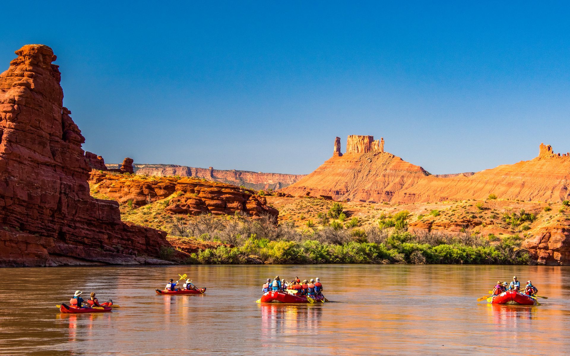 River rafters in red boats on a wide river, with red rock canyon cliffs under a blue sky.