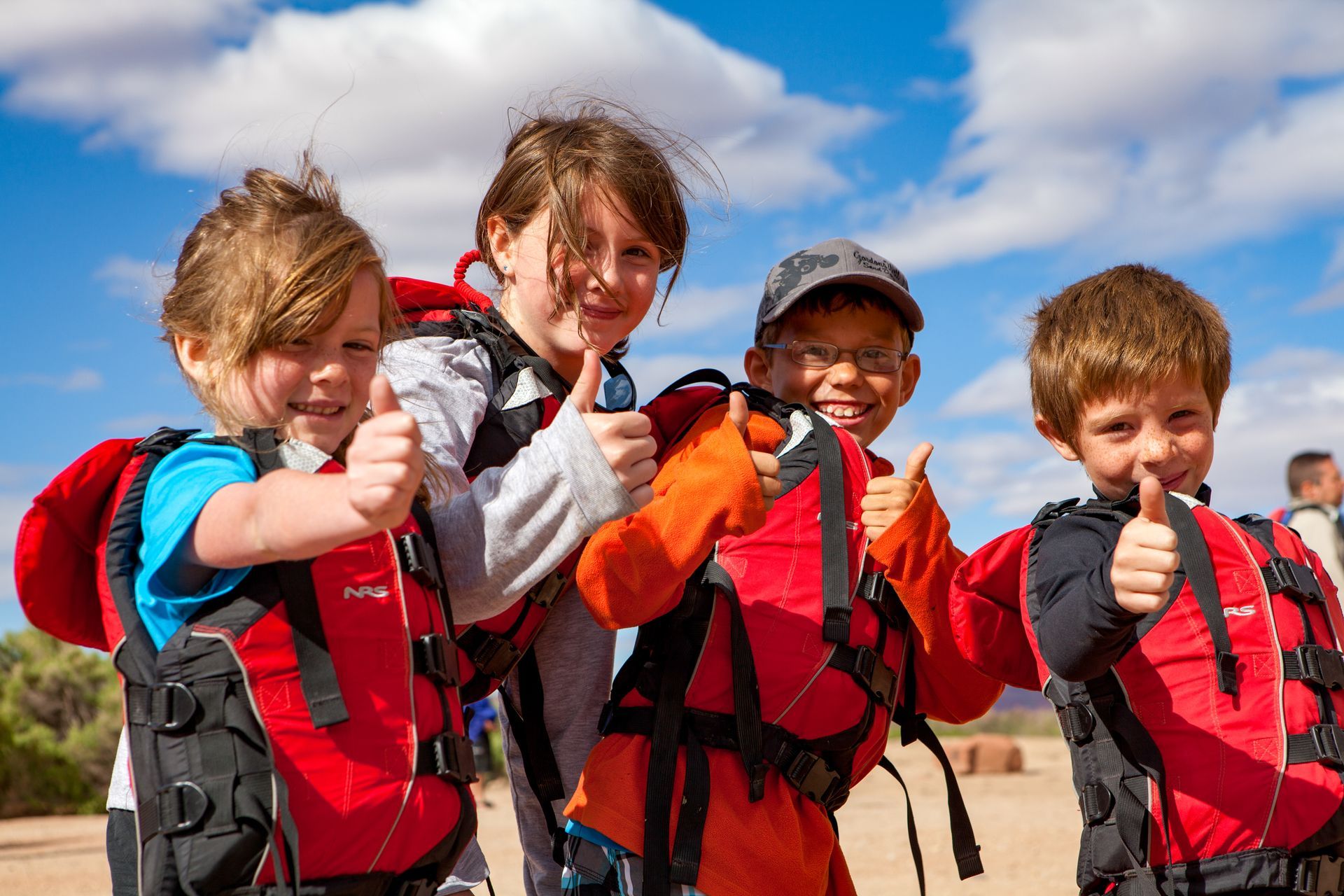 Four smiling children in life vests give thumbs up outside, under blue sky.