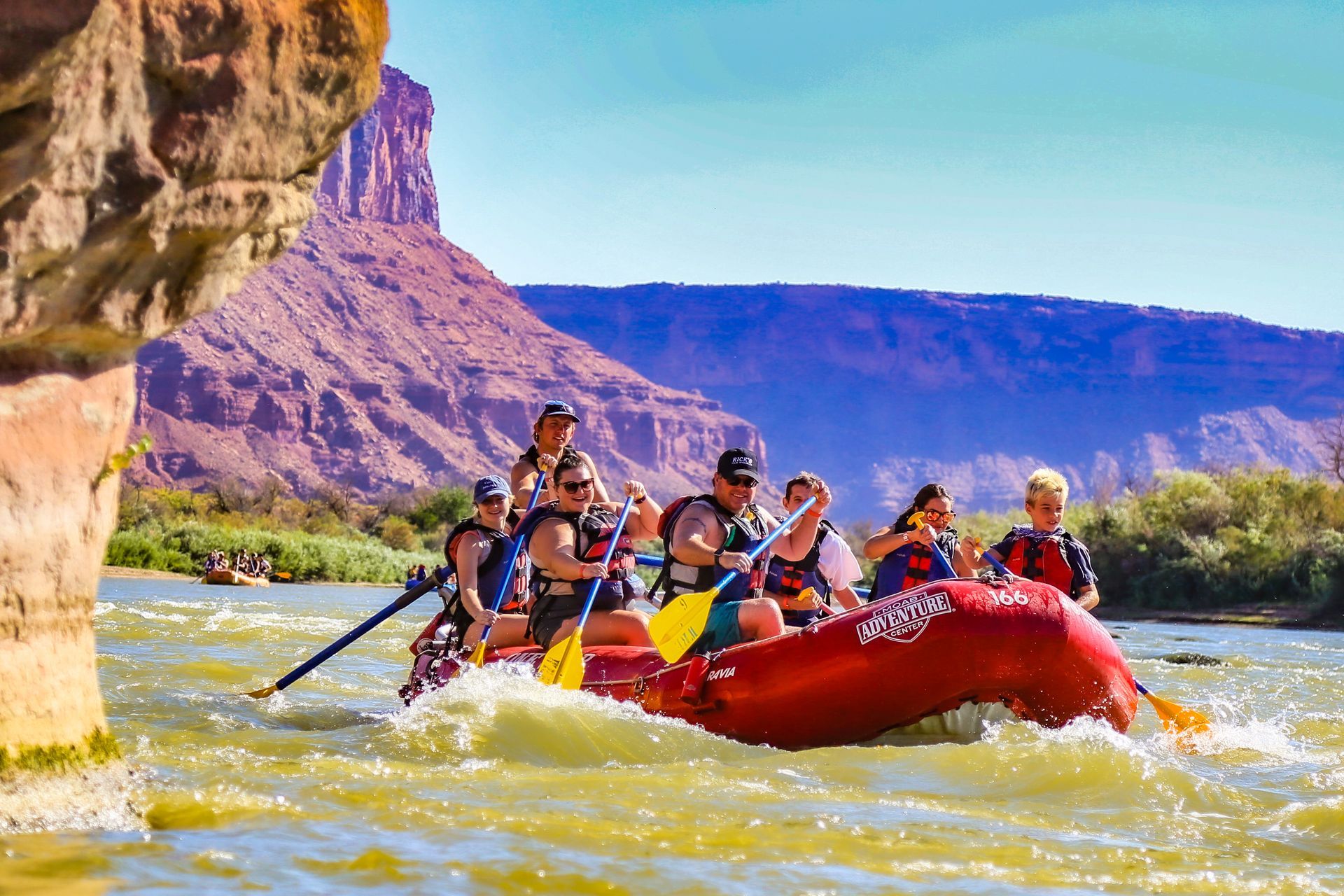 People whitewater rafting on a river, with red raft, sunny skies, and red rock canyon in the background.