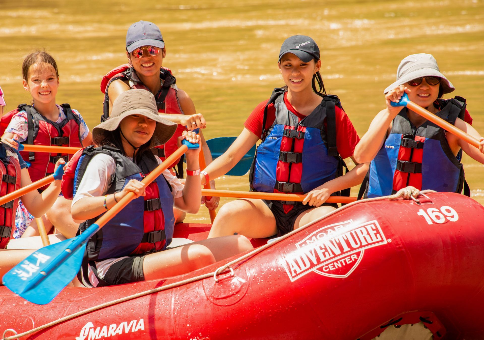 People rafting on a red boat wearing life vests; they are smiling and holding paddles in a river.