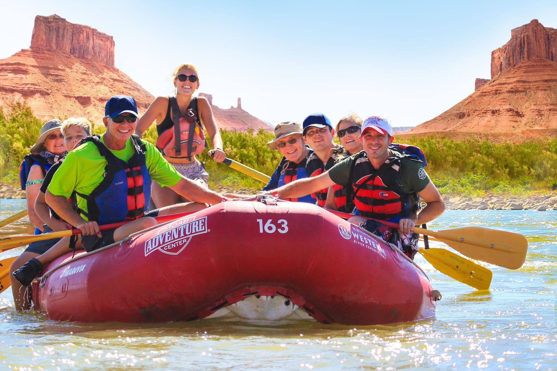 Group of people on a red raft, paddling down a river with red rock formations in the background.