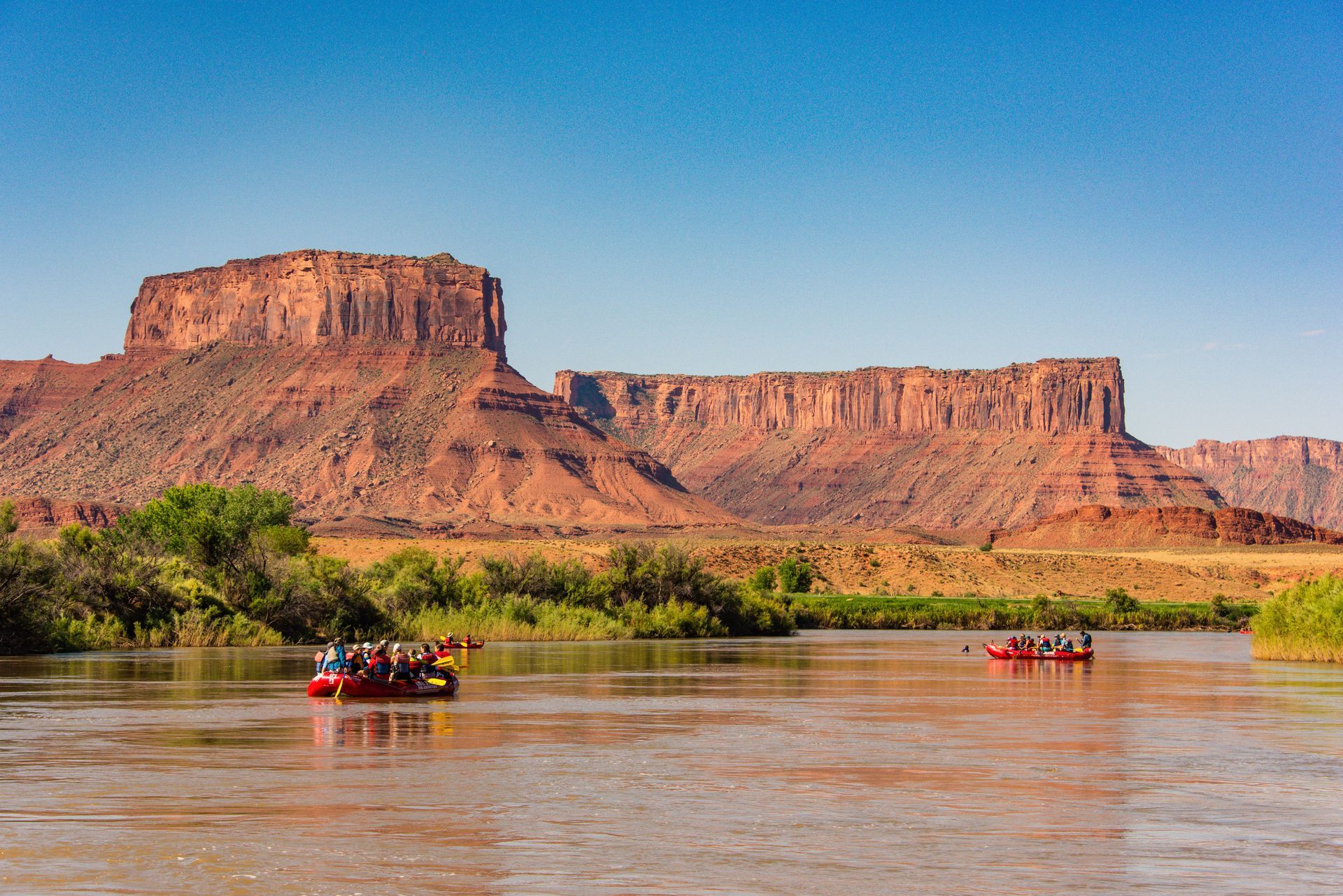 Two rafts of people float on the Colorado River with red rock cliffs in the background.