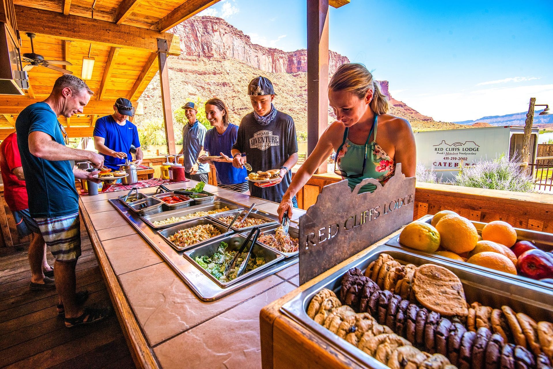 People at an outdoor buffet, selecting food. A woman selecting cookies with cliffs and a river in the background.