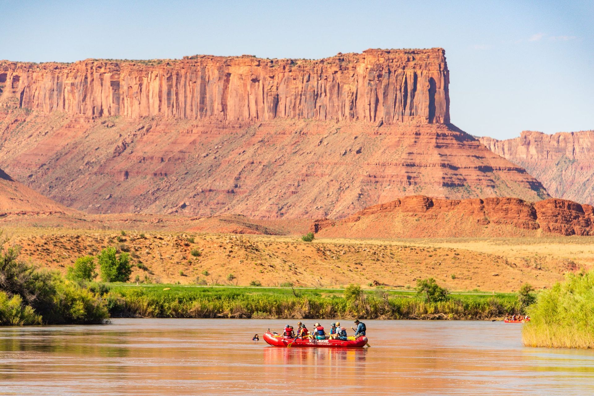Raft floating down the Colorado River on calm water with huge sandstone cliffs and green vegetation.