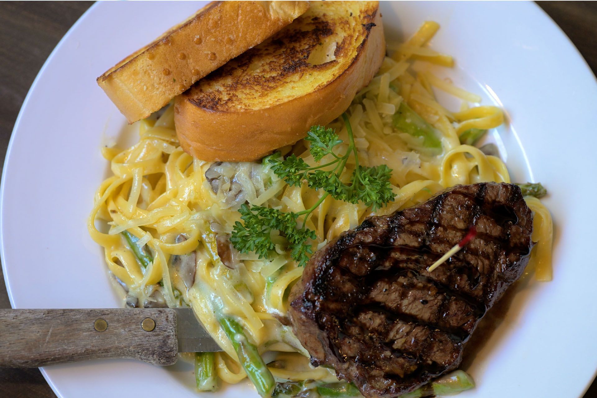 Steak with creamy pasta, asparagus, garlic bread, and a knife on a white plate.