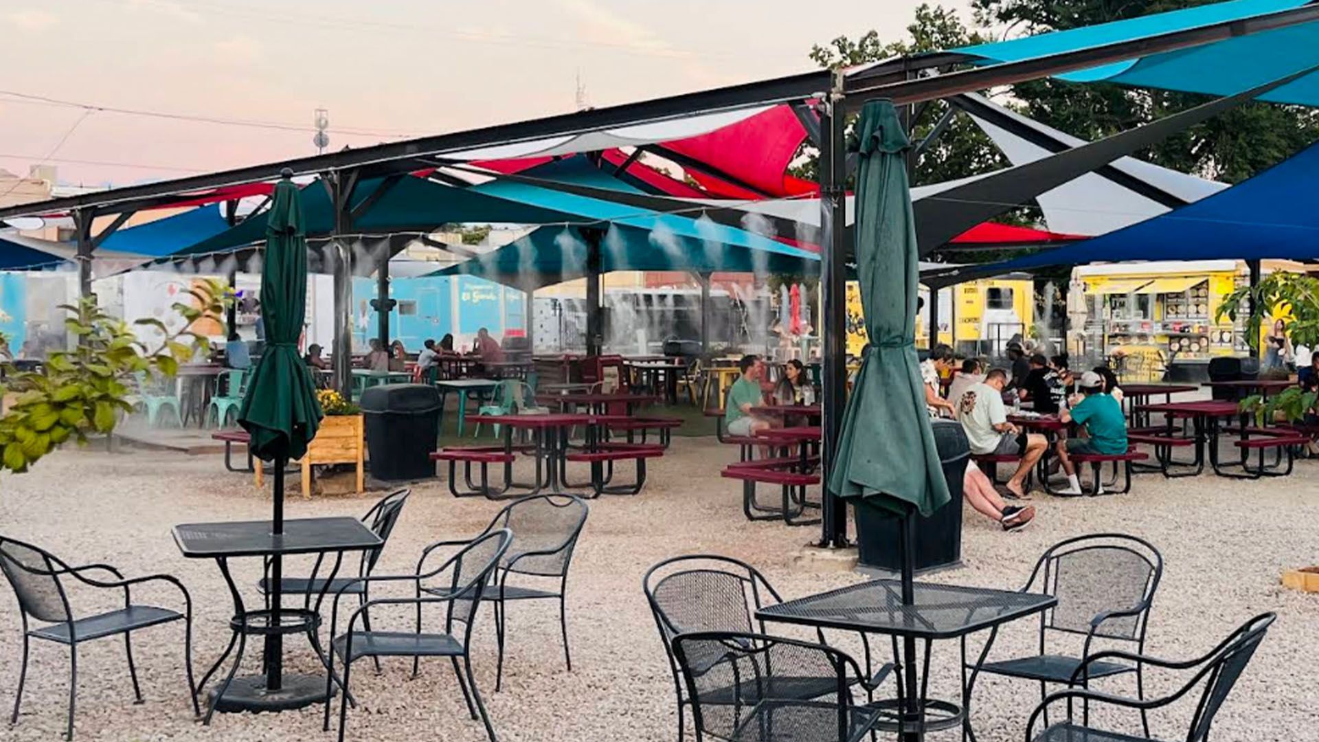 Outdoor dining area with tables, umbrellas, and colorful shade sails; people are seated and socializing.