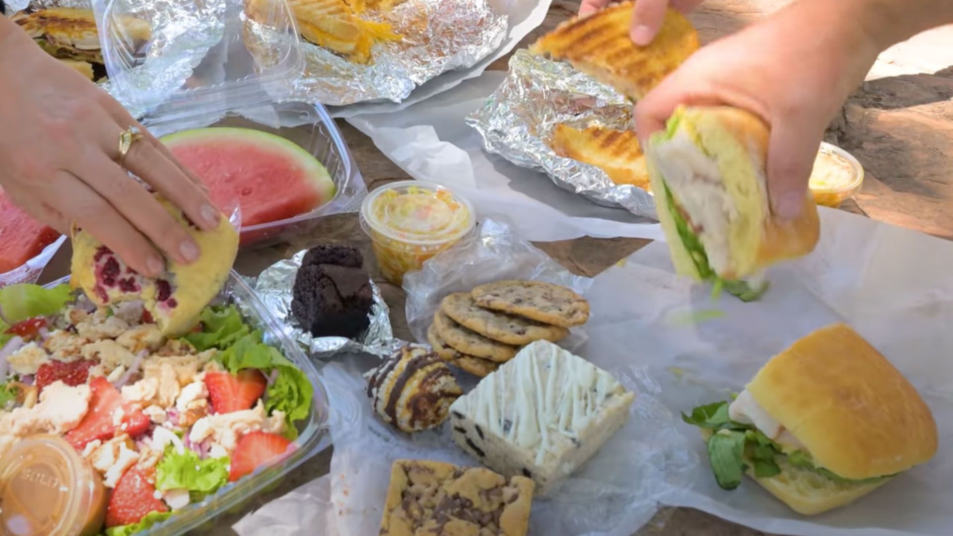 Hands reaching for picnic food including salad, sandwiches, fruit, and desserts on a table.