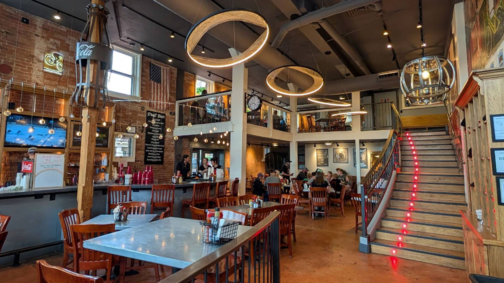 Interior of a restaurant with tables, stairs, bar, and multiple levels. Exposed brick, American flag, and hanging lights.