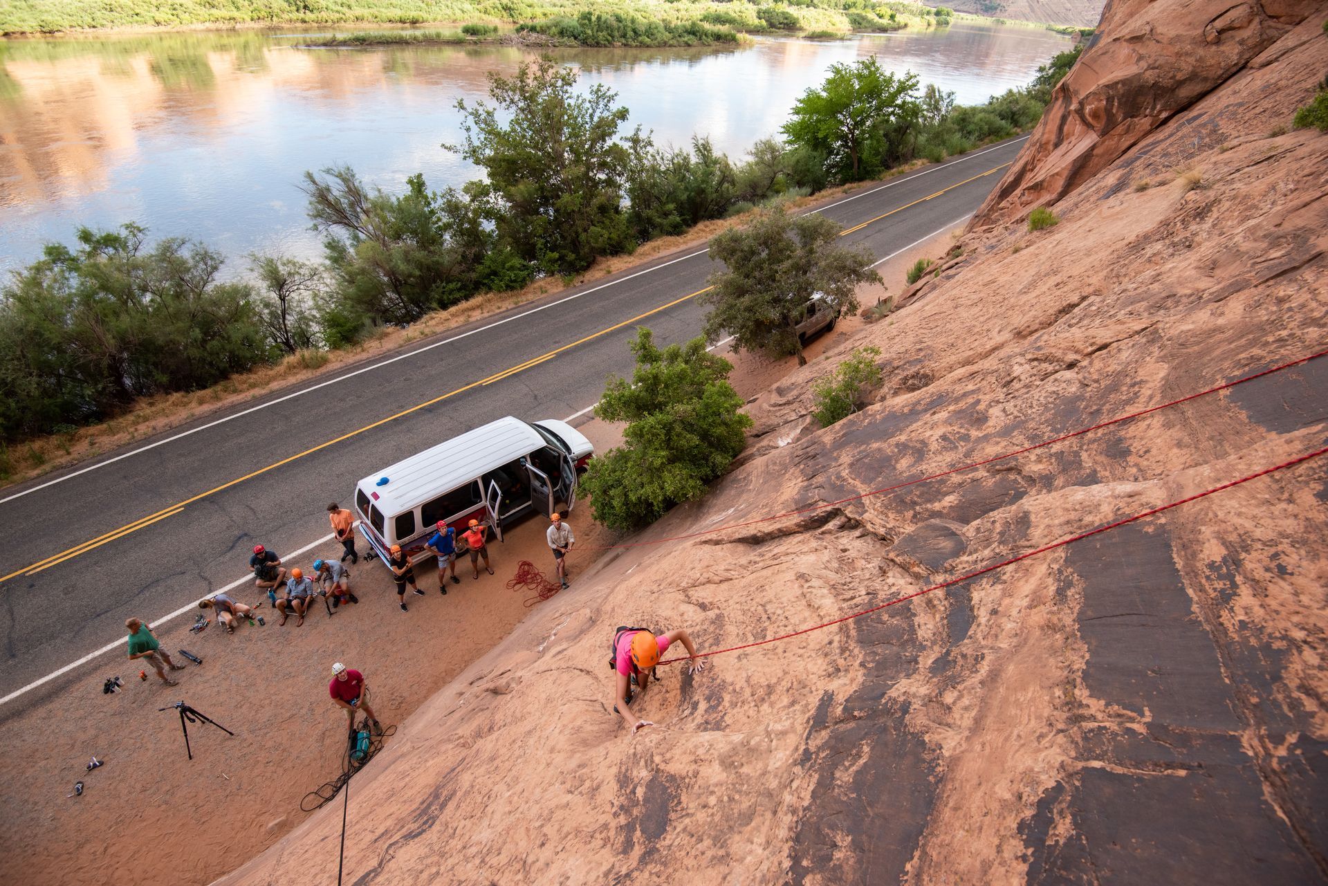 Rock climbers near a road, van, and river. One person climbs the rock face.