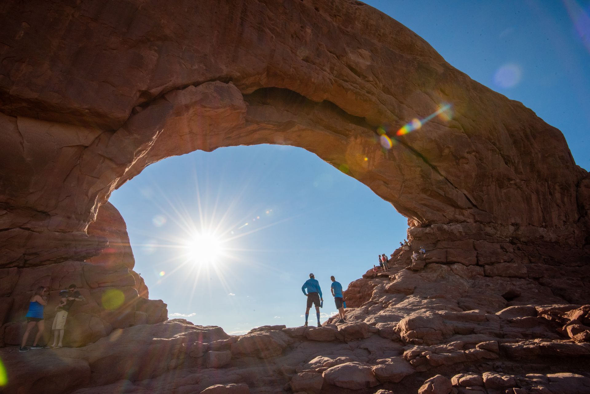 Two people are standing under North Window Arch in Arches National Park with the sun shining through the Arch.