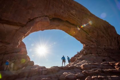 Two people stand under a massive stone arch with the sun shining through the middle.