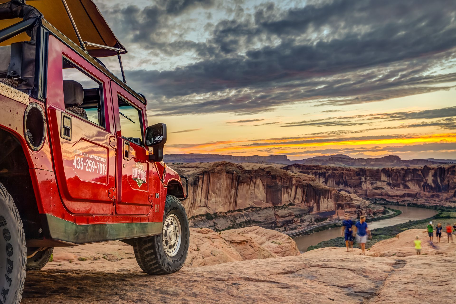 Red off-road Hummer on a rocky trail at sunset, with hikers below and canyon cliffs in the background.