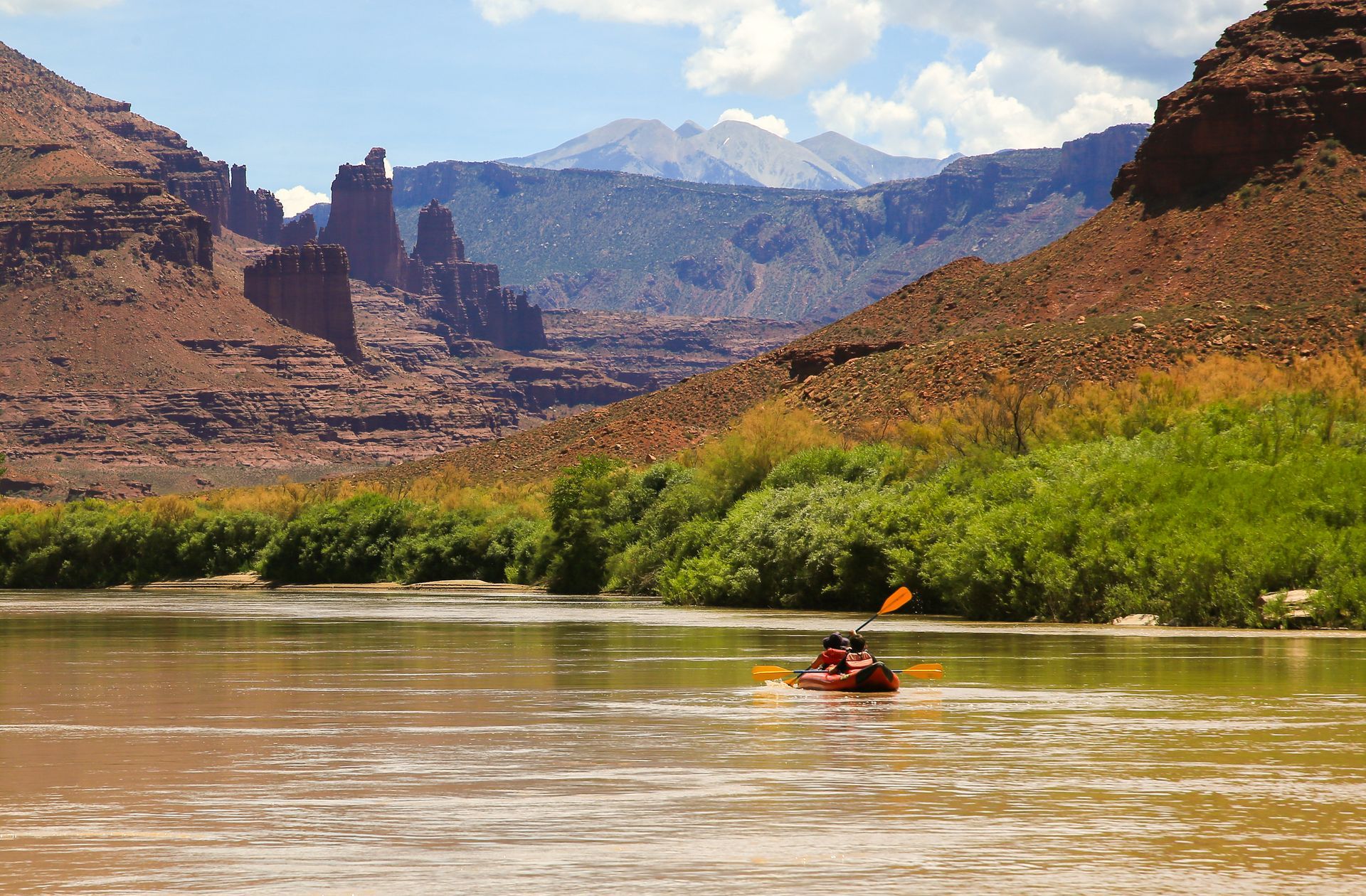 Kayakers paddling downriver navigate past green foliage and red rock formations with a mountain in the distance.