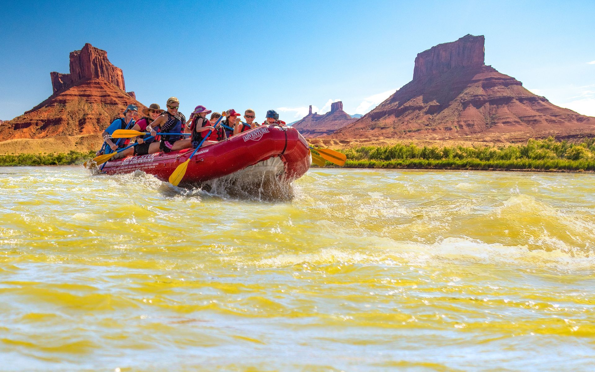 Red raft with people paddling on a river, passing red rock formations under a blue sky.