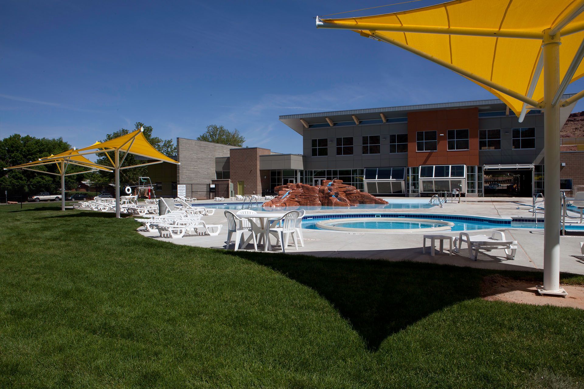 Pool area with yellow shade sails, white chairs, a building and a grassy lawn.