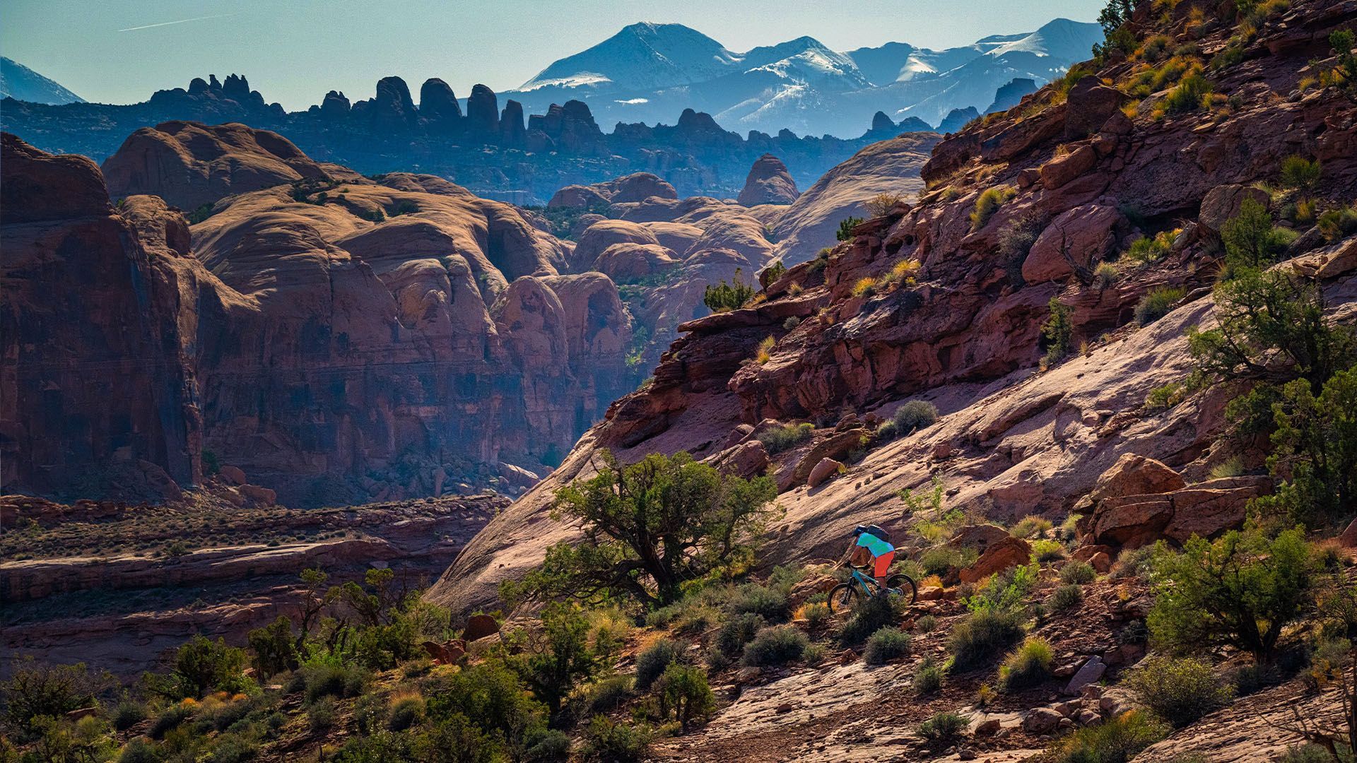Rugged canyon landscape with hiker in bright clothing; sunlit rocks, distant mountains.
