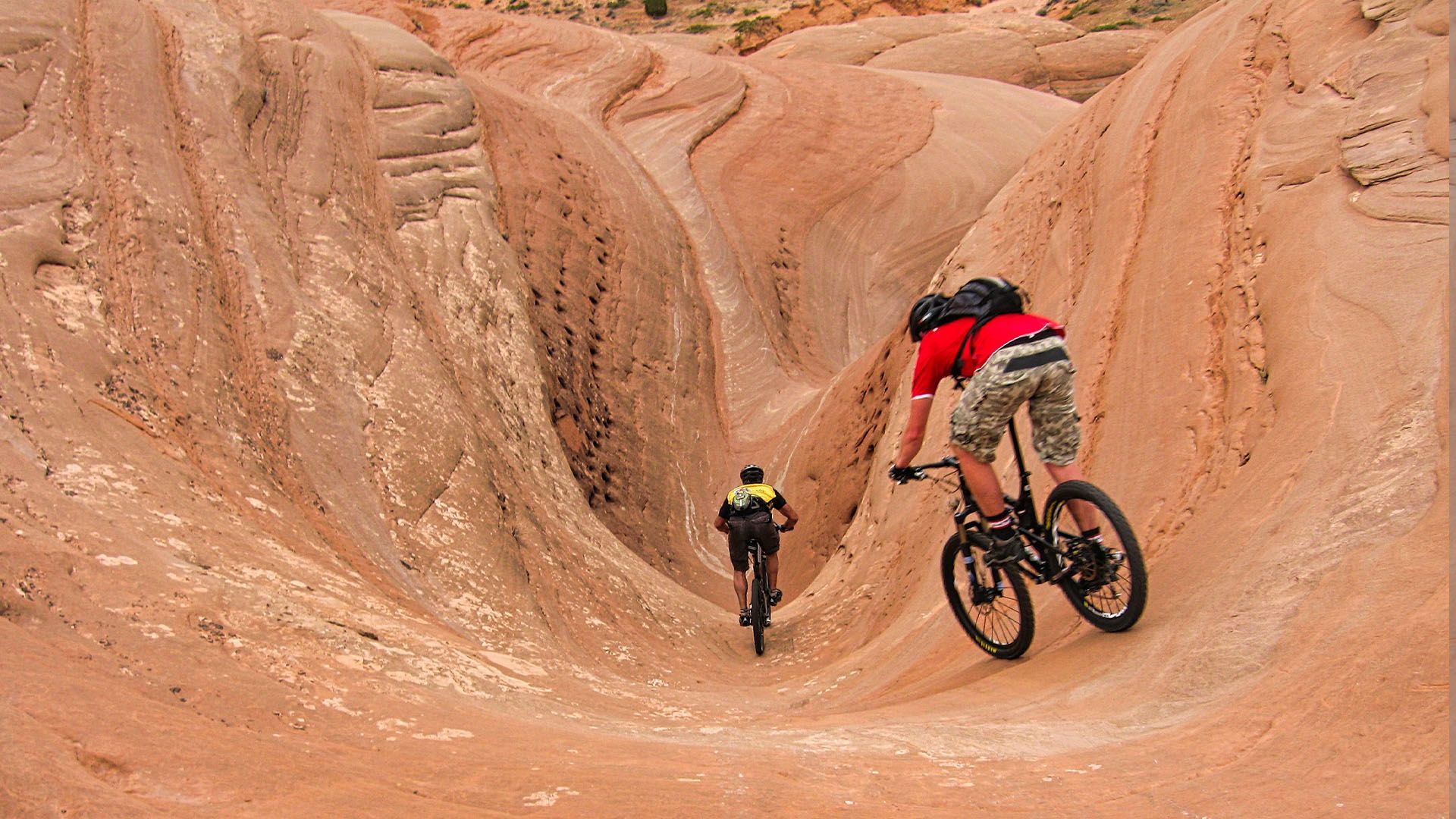 Two mountain bikers riding down a sandy, eroded canyon trail. Red rock, sunny day.