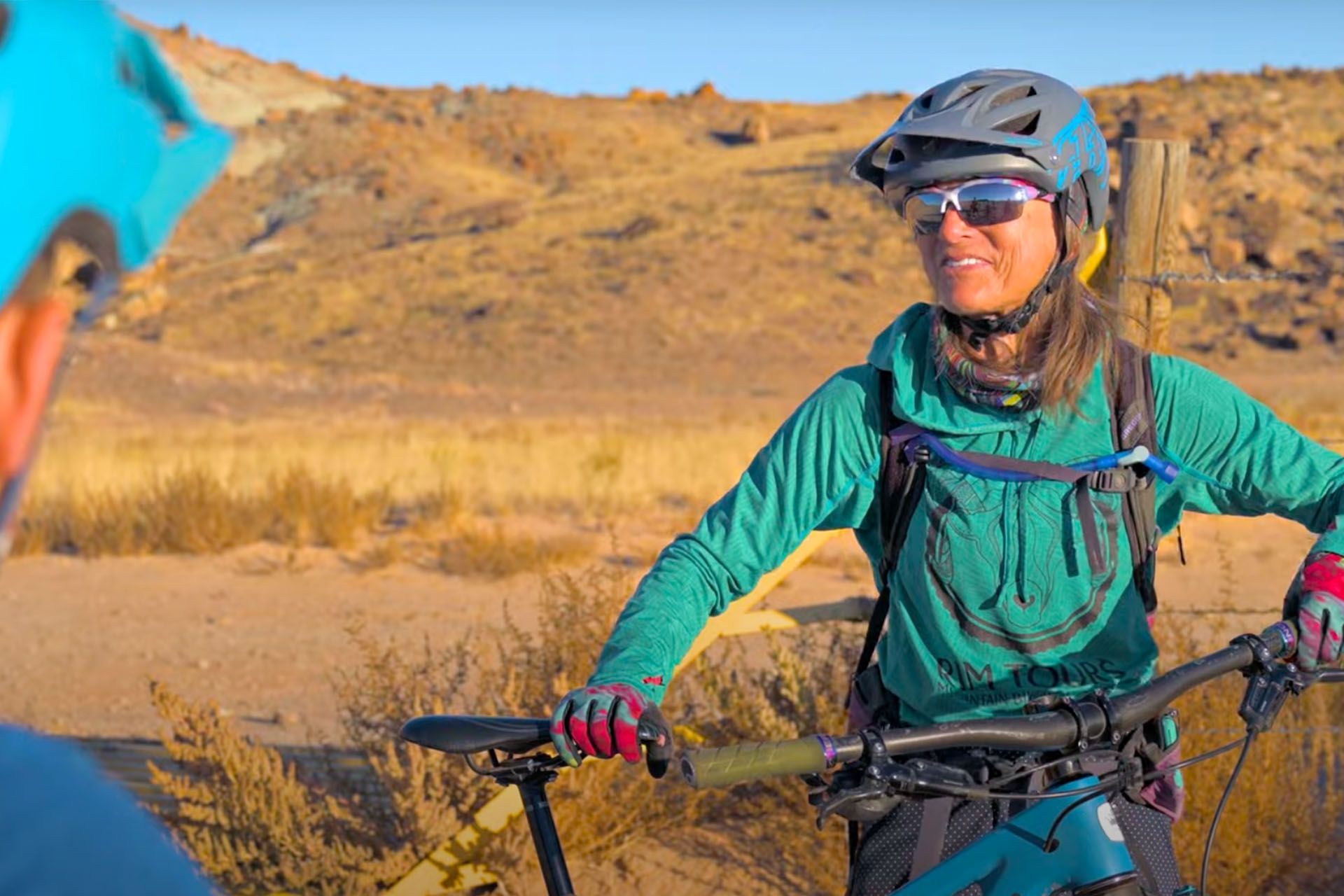 Woman on a mountain bike smiles, wearing a helmet and sunglasses, with a desert landscape in the background.