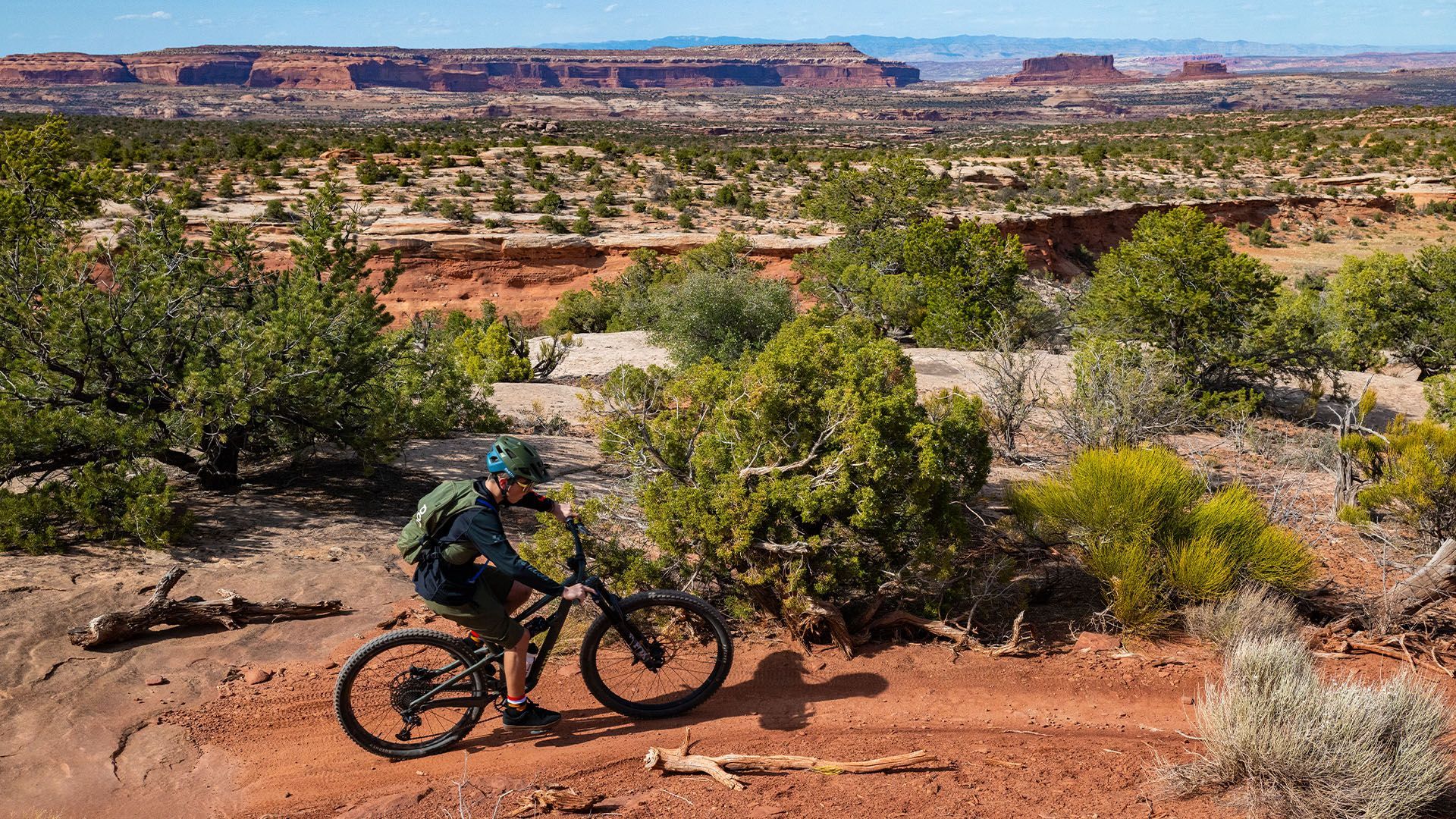 Mountain biker riding on a red dirt trail with desert landscape and mesas in the background.