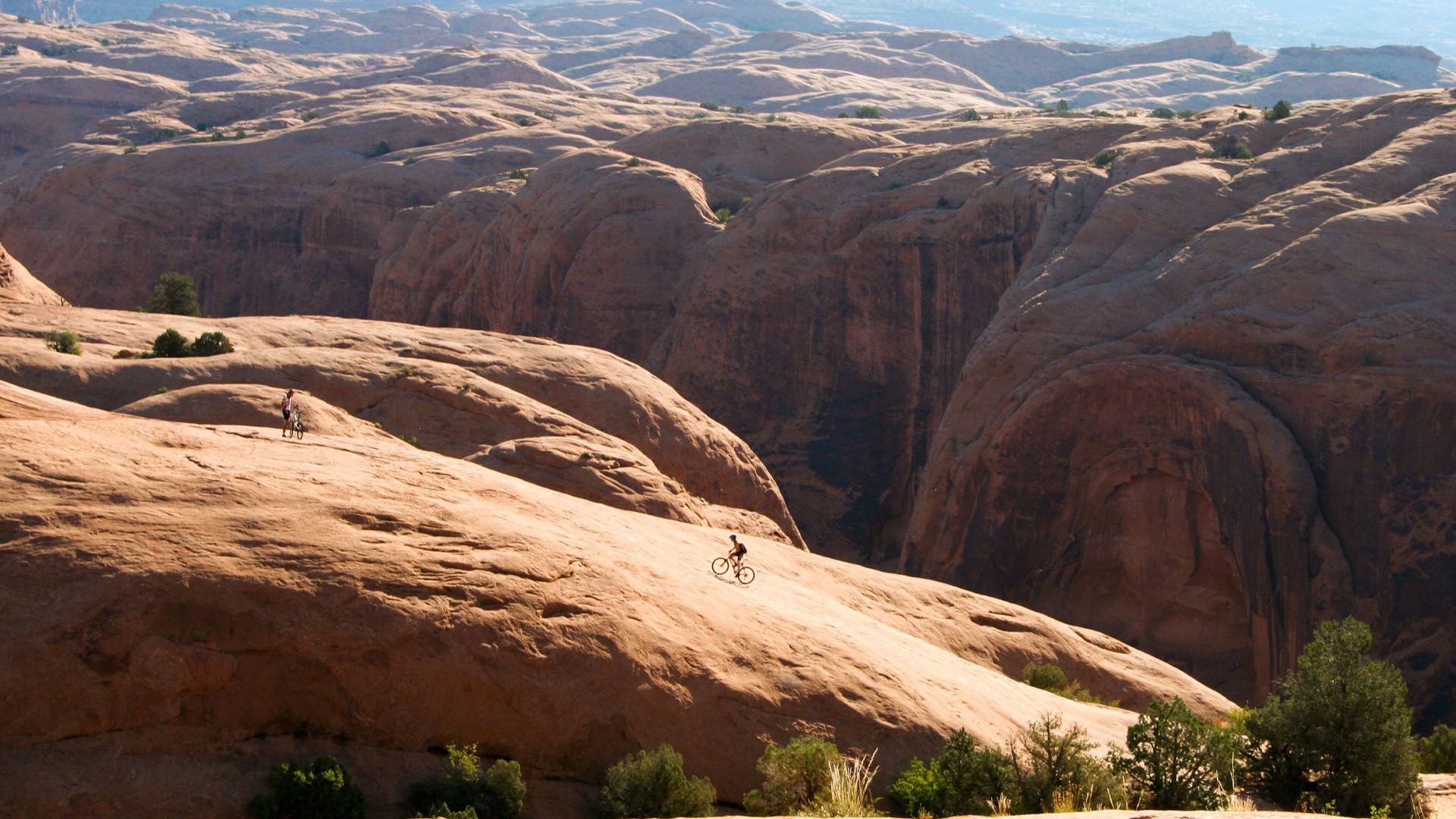 A cyclist rides across a large, tan rock formation with a canyon in the distance; sunny landscape.