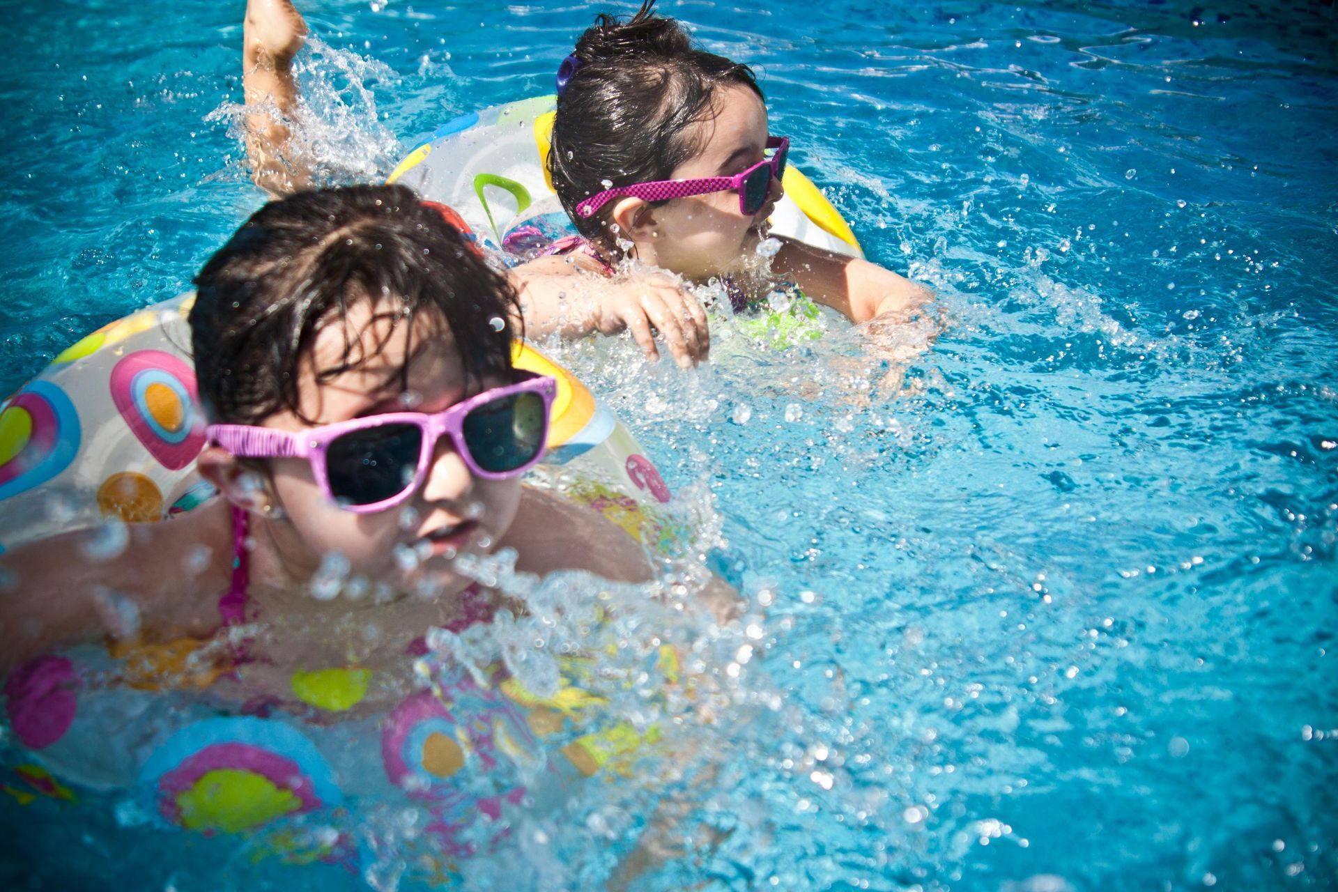 Two girls wearing sunglasses in a pool, one with an inflatable ring, splashing and smiling.