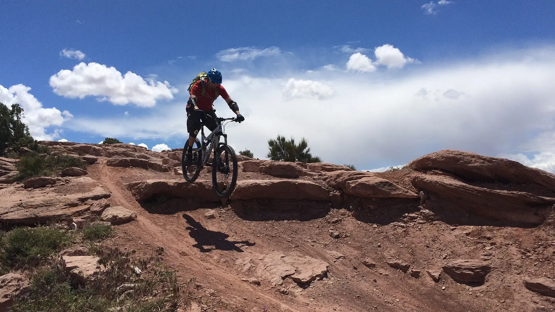 Mountain biker riding downhill on a rocky red trail under a blue sky with fluffy clouds.