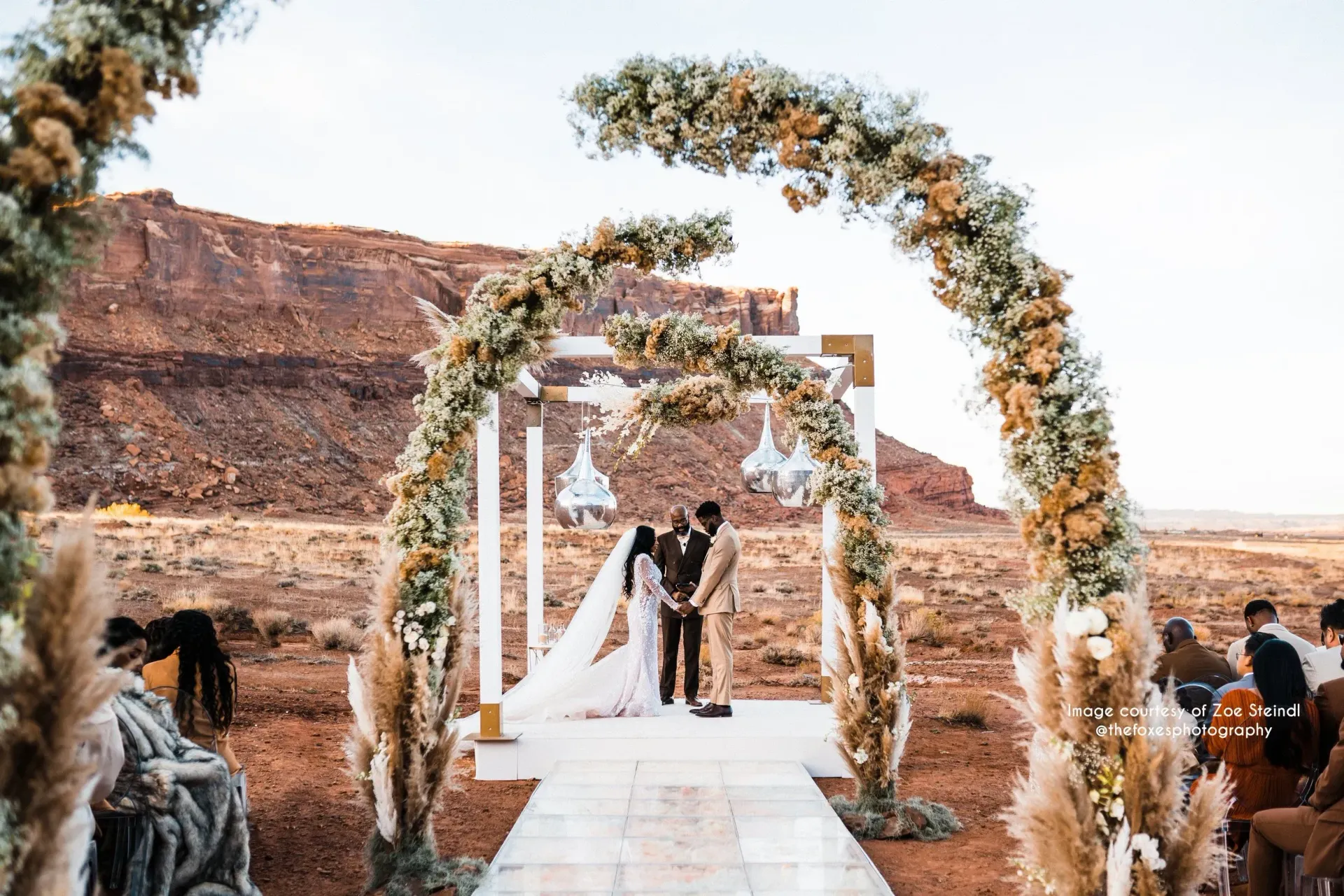 Couple exchanging vows under a floral arch in a desert setting.