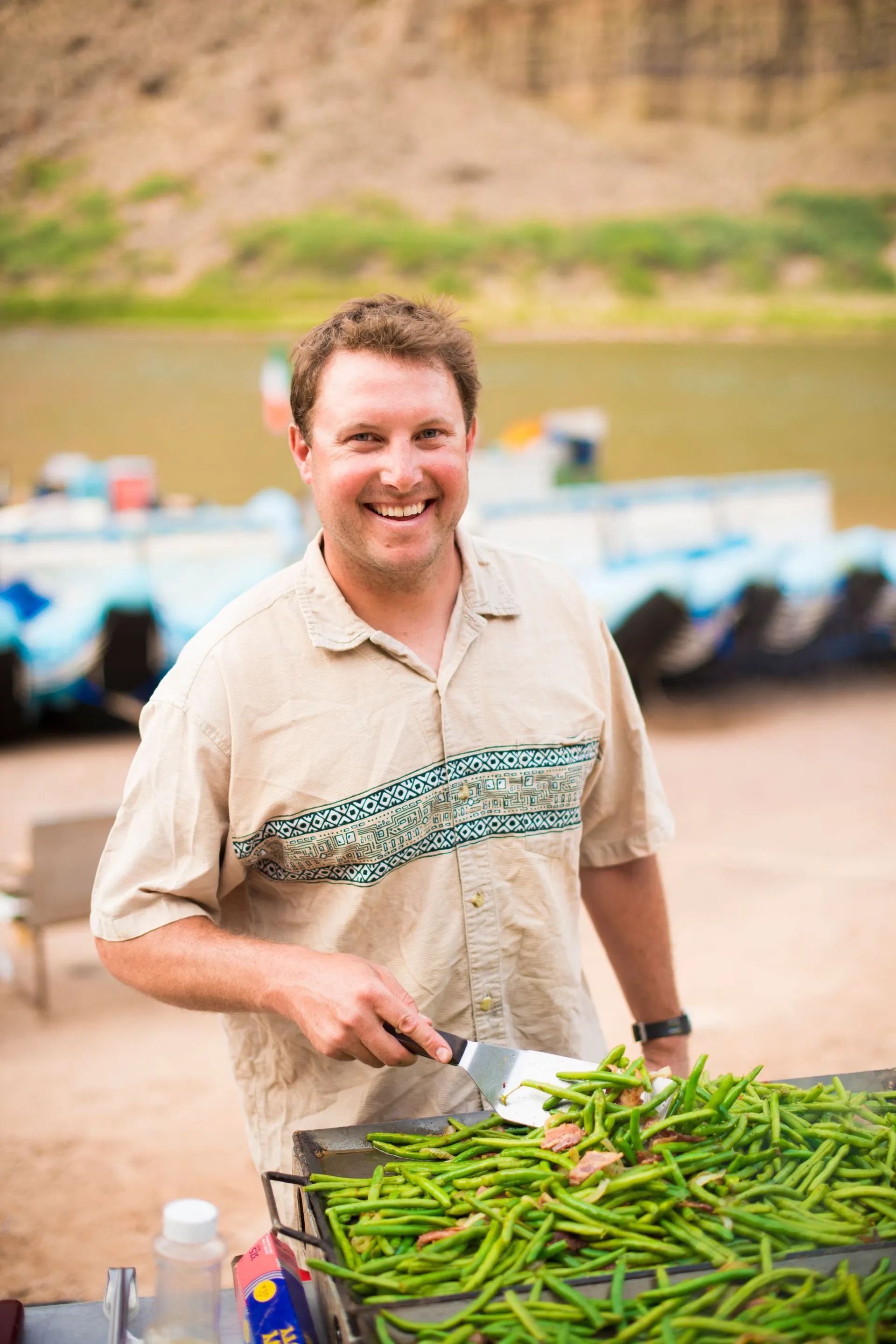 Man cooking green beans on a grill outdoors, smiling. River and boats in background.