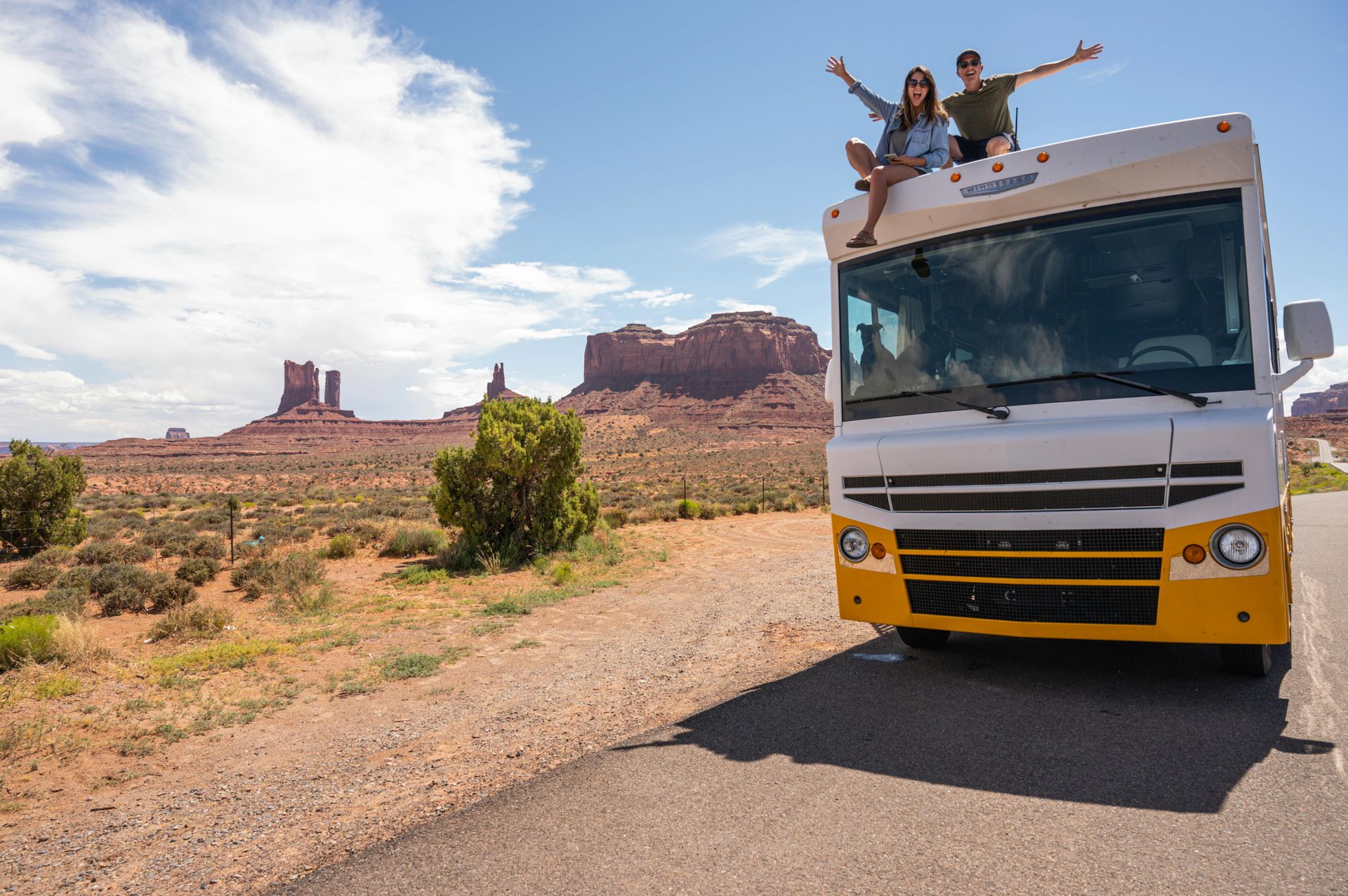 Couple atop an RV with arms raised, enjoying scenic desert landscape under a blue sky.