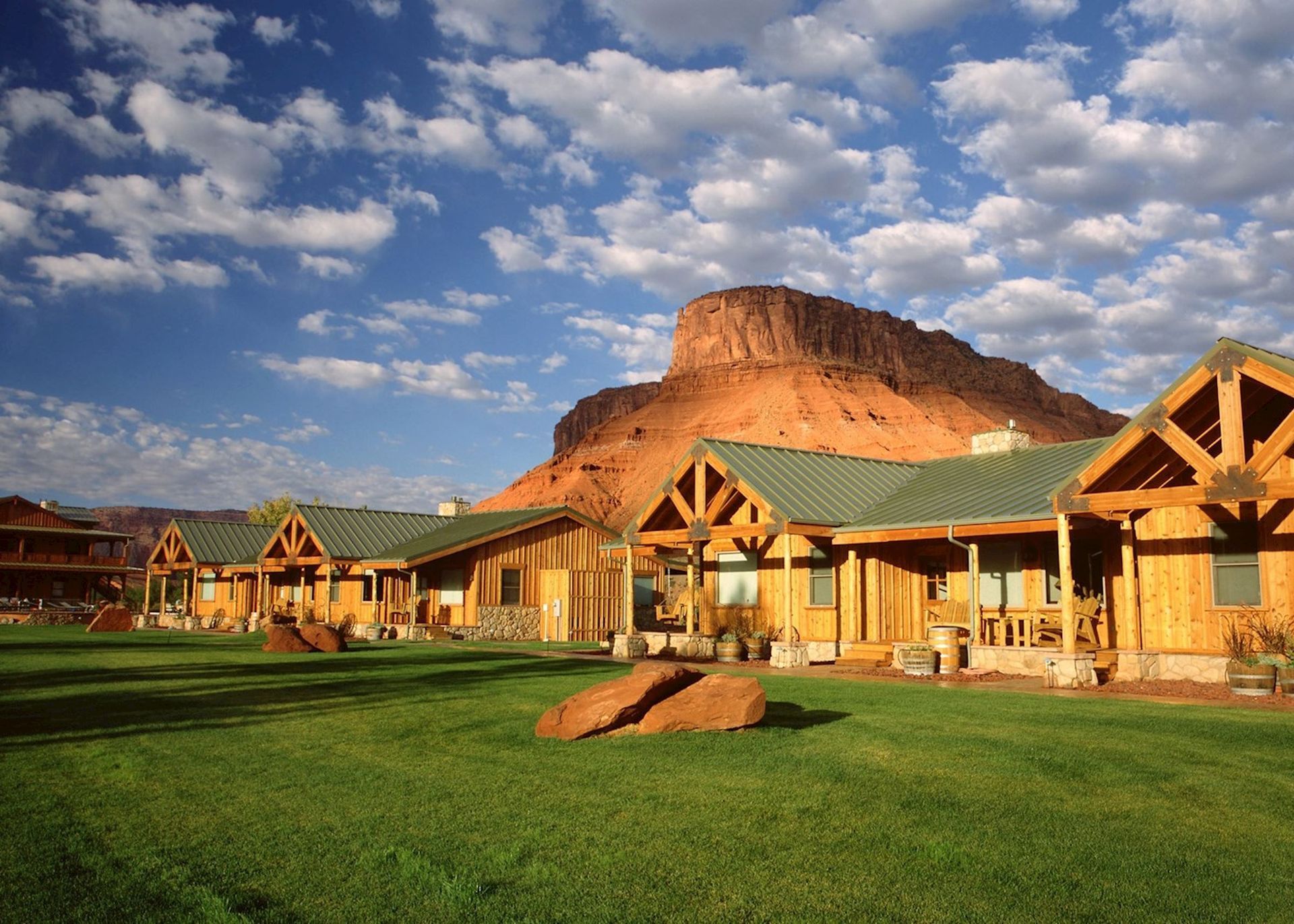 Wooden cabins with green roofs and a grassy lawn, with a large red rock mountain and blue sky.