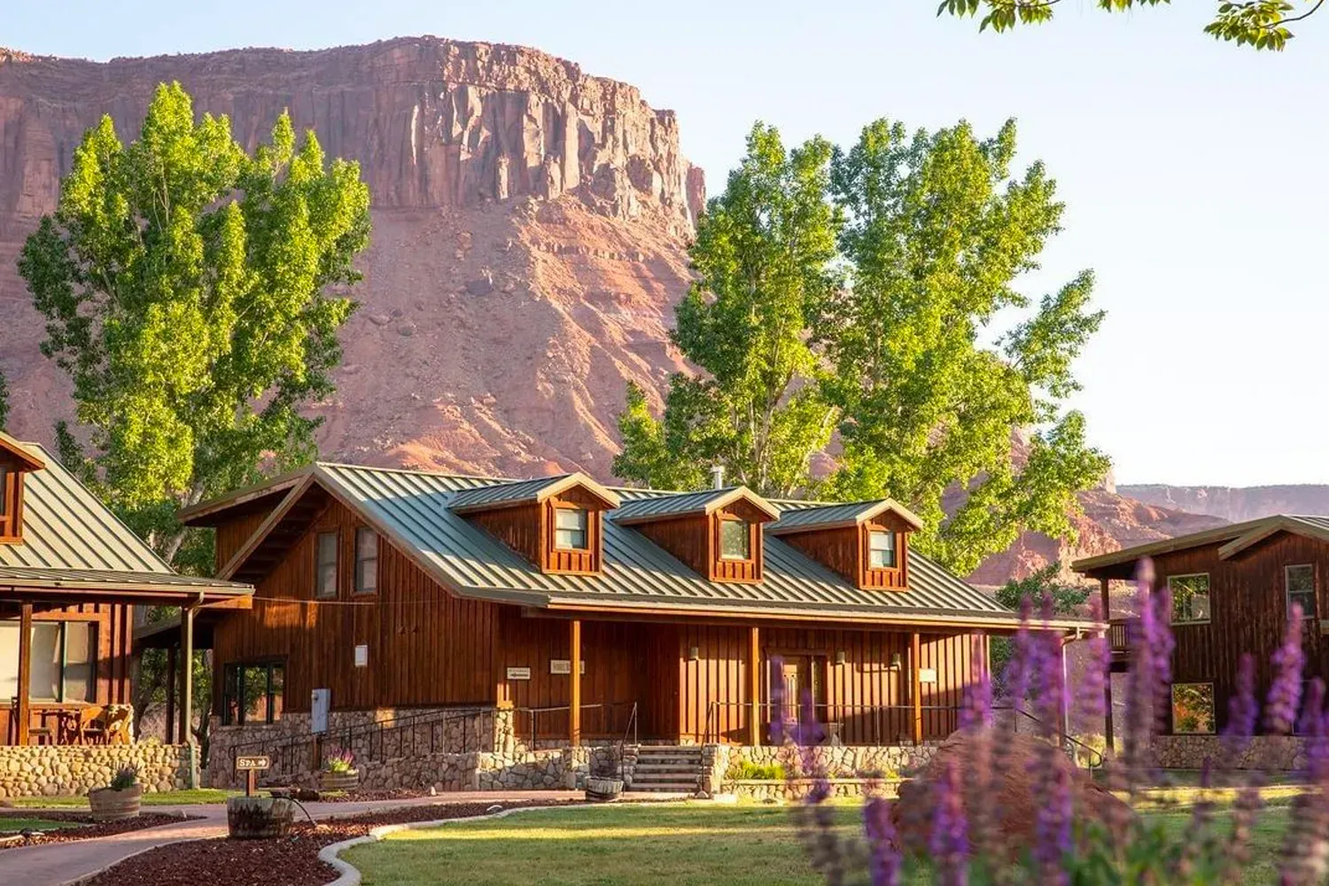 Wooden cabins with green trees and purple flowers, red rock mountains in the background.