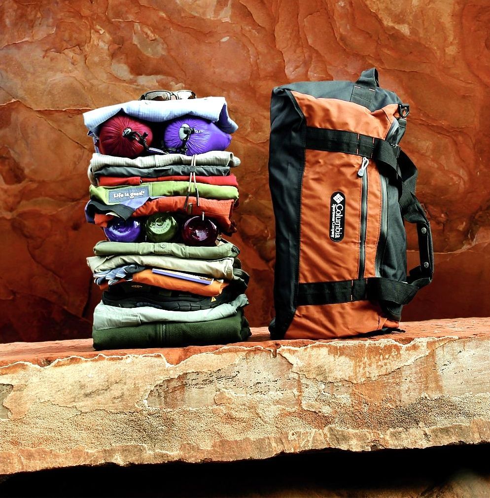 Stack of colorful clothing and gear beside an orange and black duffel bag on a stone ledge.