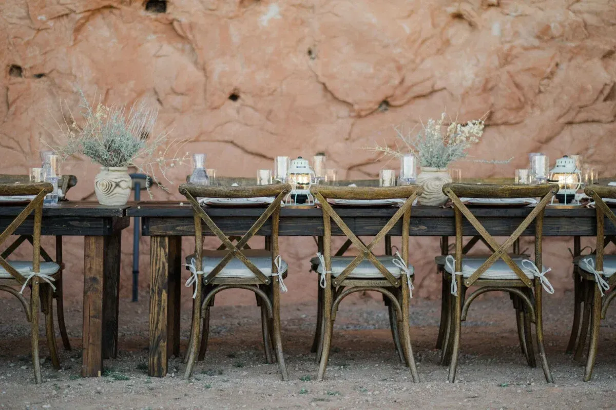 Wooden tables and chairs set for a wedding reception against a rock wall.