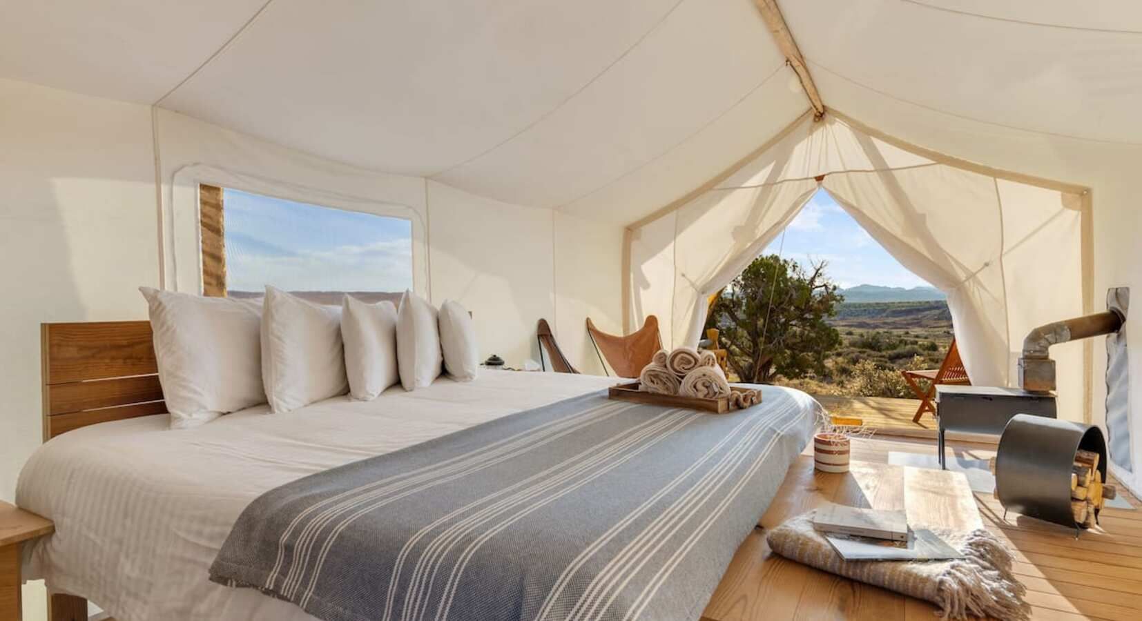 Luxurious glamping tent interior: bed, pillows, wood-burning stove, landscape view.