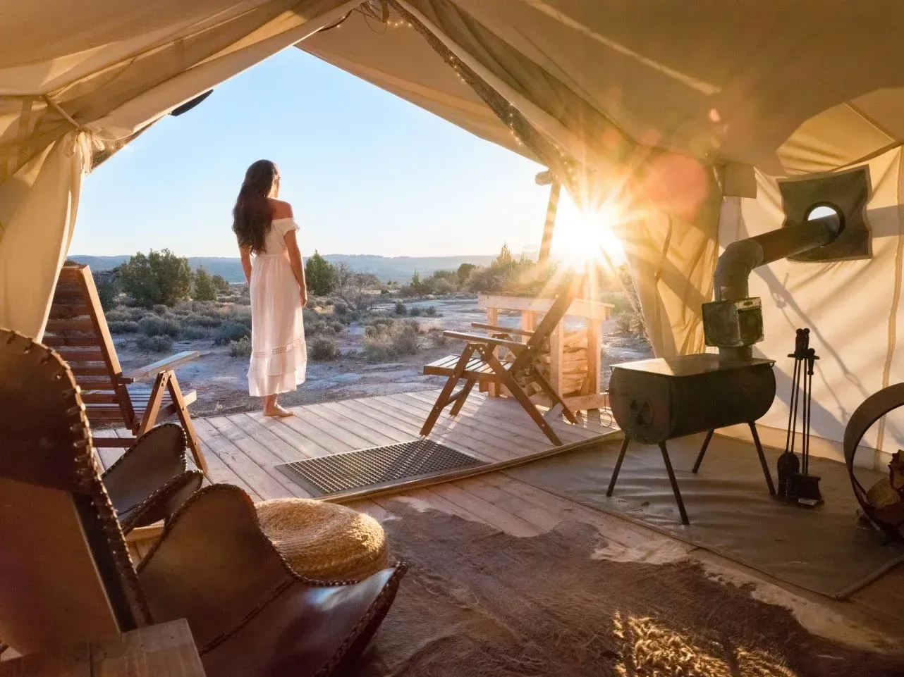 Woman in white dress stands at glamping tent entrance, watching sunrise over desert landscape.